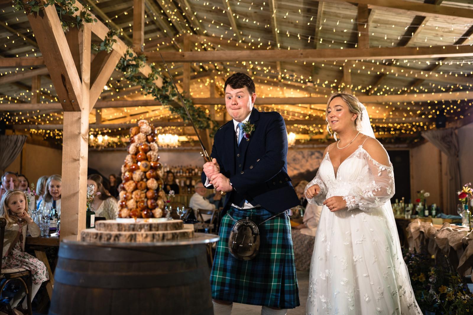 Groom Hamish makes a funny, determined face as he uses a large sword to cut their unique croquembouche wedding cake on a whiskey barrel while bride Emma watches on.