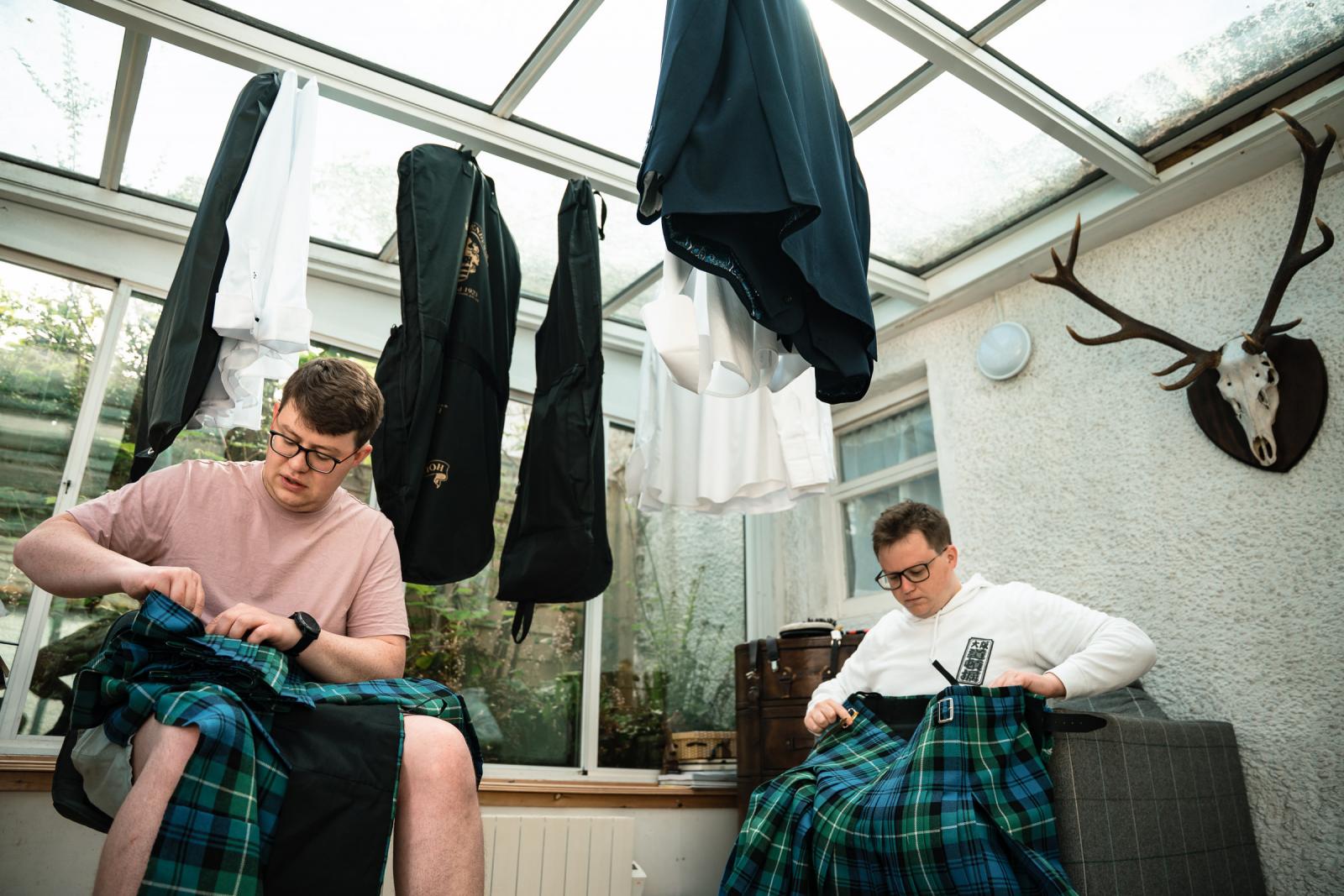 Hamish and his groomsman sitting in a bright sunroom at home, preparing their traditional Scottish blue and green tartan kilts during the wedding morning preparations.