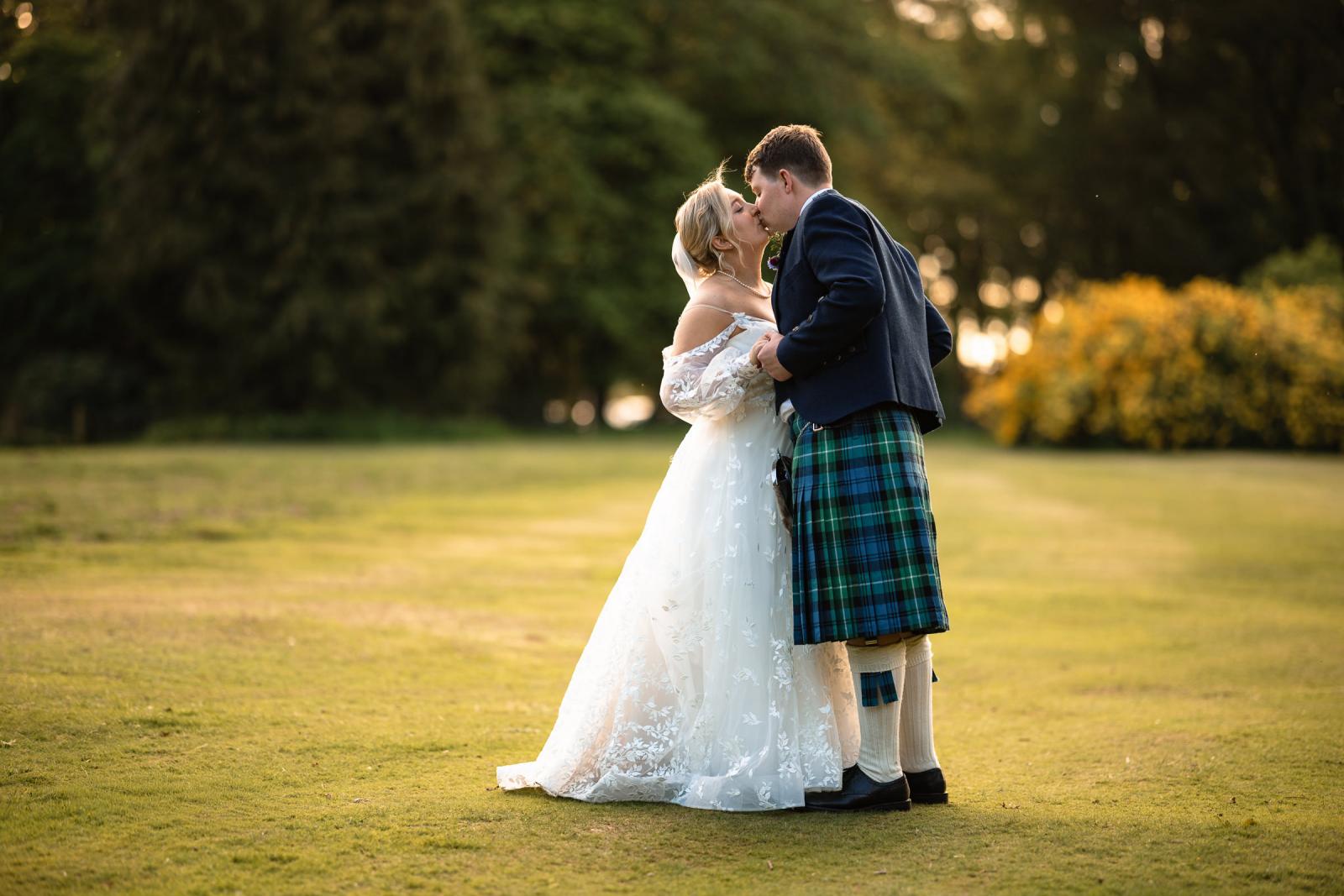 Romantic golden hour wedding portrait of Emma and Hamish sharing a sweet kiss on a vast green lawn at sunset, surrounded by beautiful Scottish woodlands.