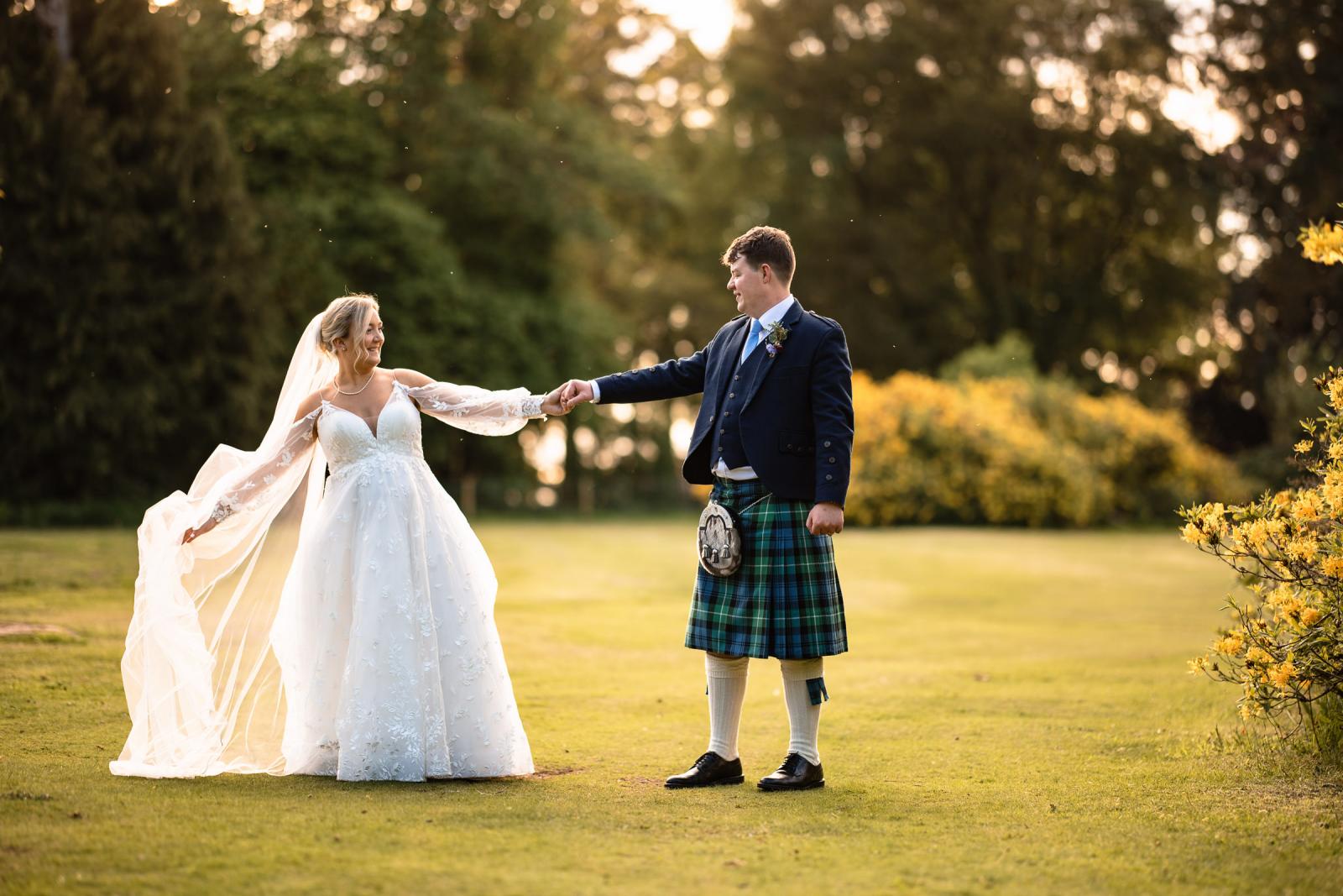 Full-length portrait of Emma and Hamish holding hands on a sun-drenched lawn at Culdees Castle Estate, with the groom looking lovingly at his bride during golden hour.
