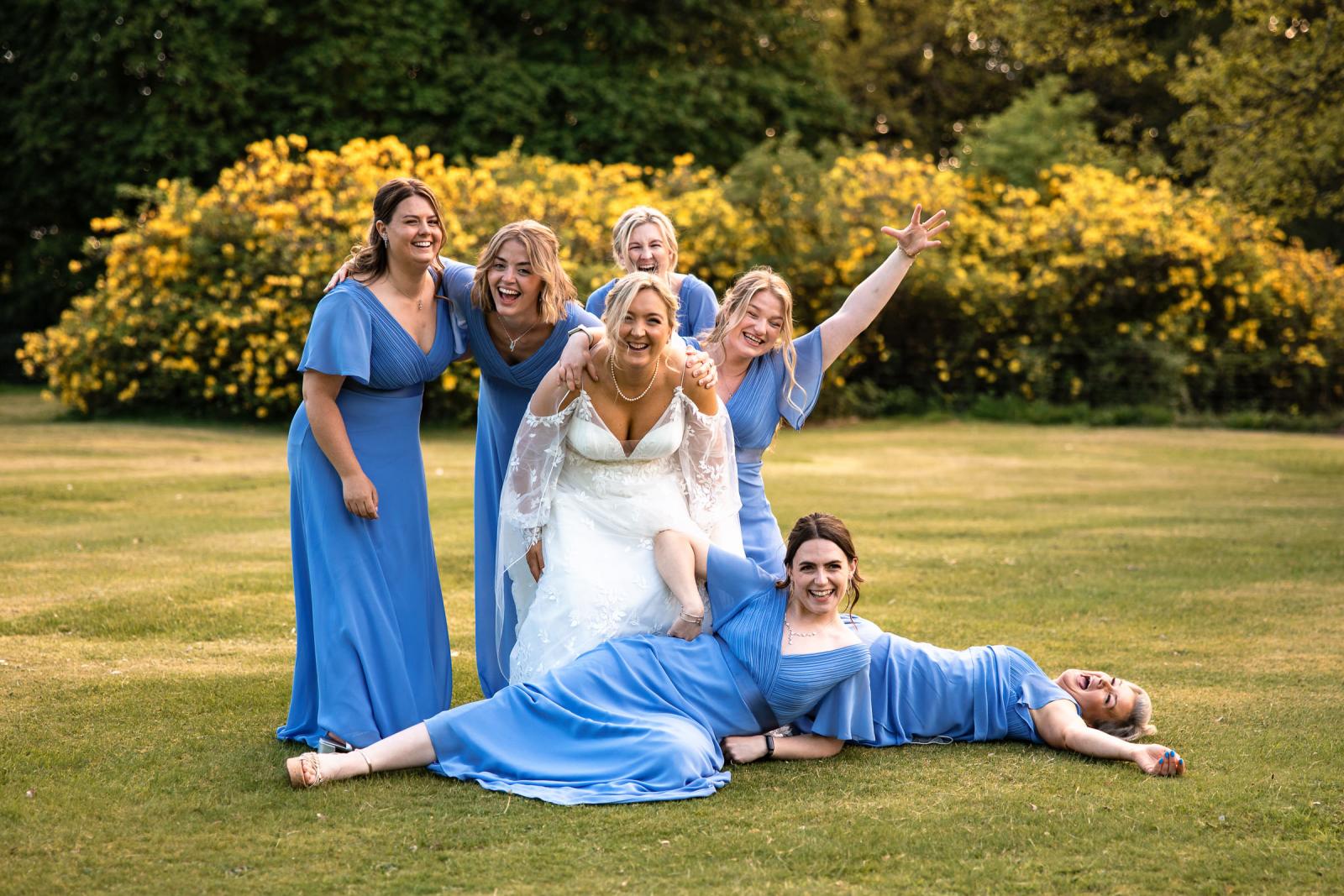 A playful group photo of bride Emma laughing with her bridesmaids in cornflower blue dresses, with two bridesmaids comically posing on the grass.