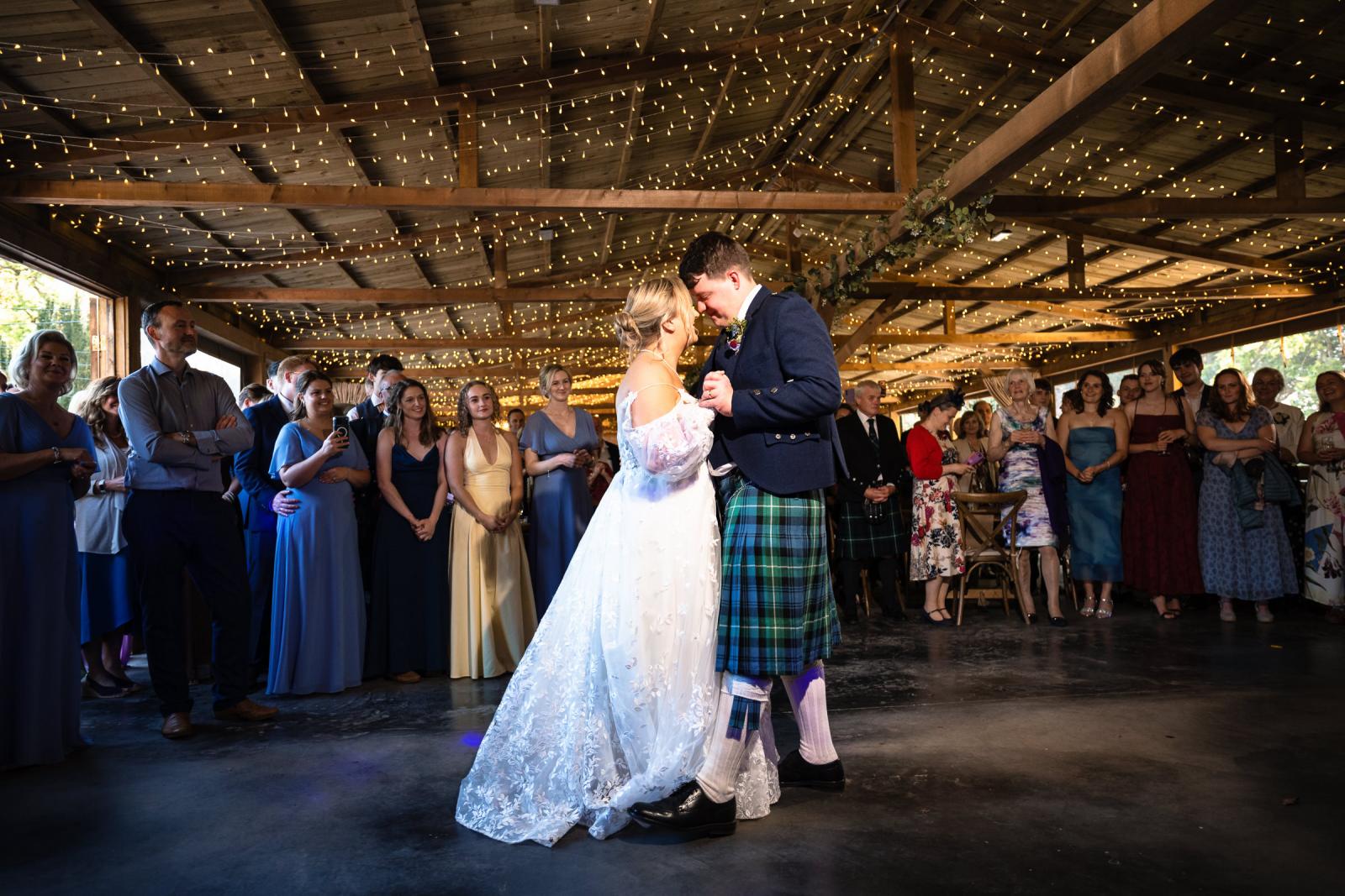 Emma and Hamish holding each other close during their romantic first dance inside the barn, beneath a canopy of twinkling fairy lights as wedding guests look on.