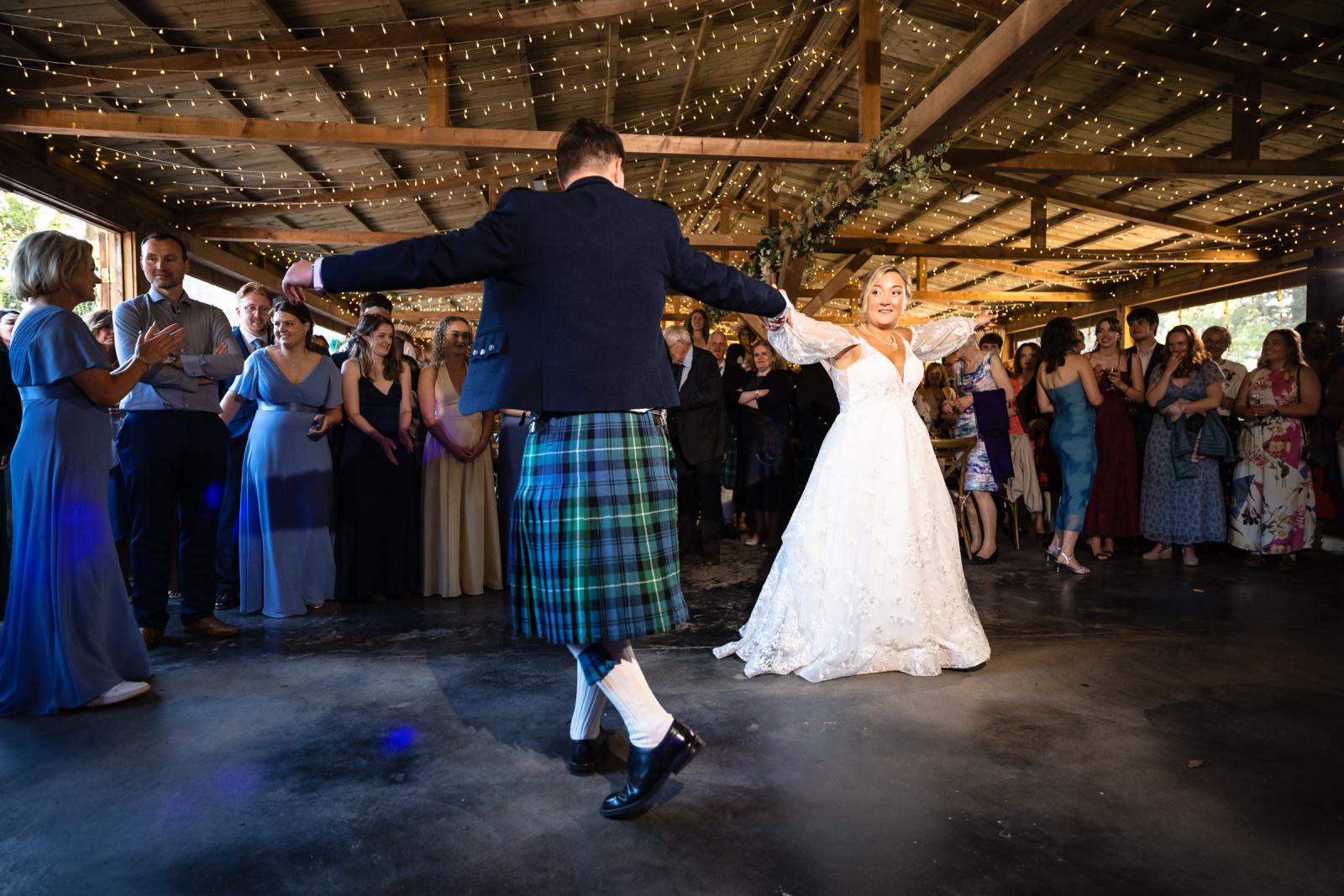 Groom Hamish spins around with arms wide open during the first dance, showing off his traditional Scottish kilt as bride Emma smiles.