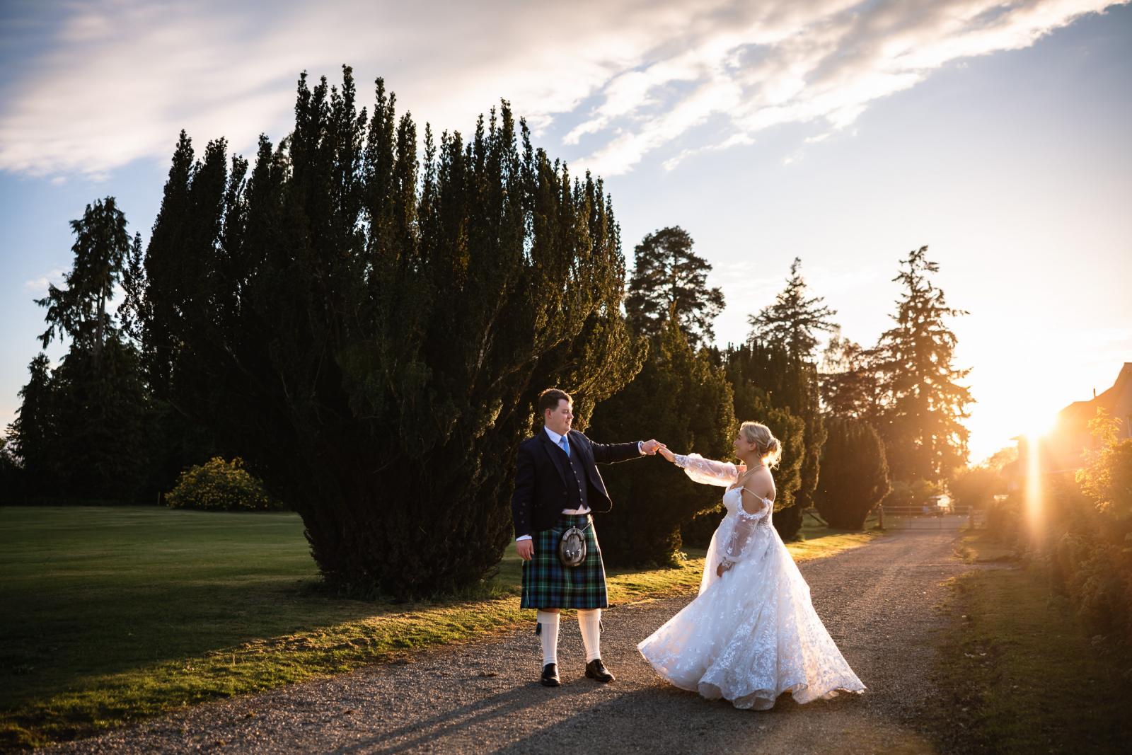 The newlyweds playfully twirl on a gravel path during sunset, with a beautiful sun flare bursting through the tall trees in the background.