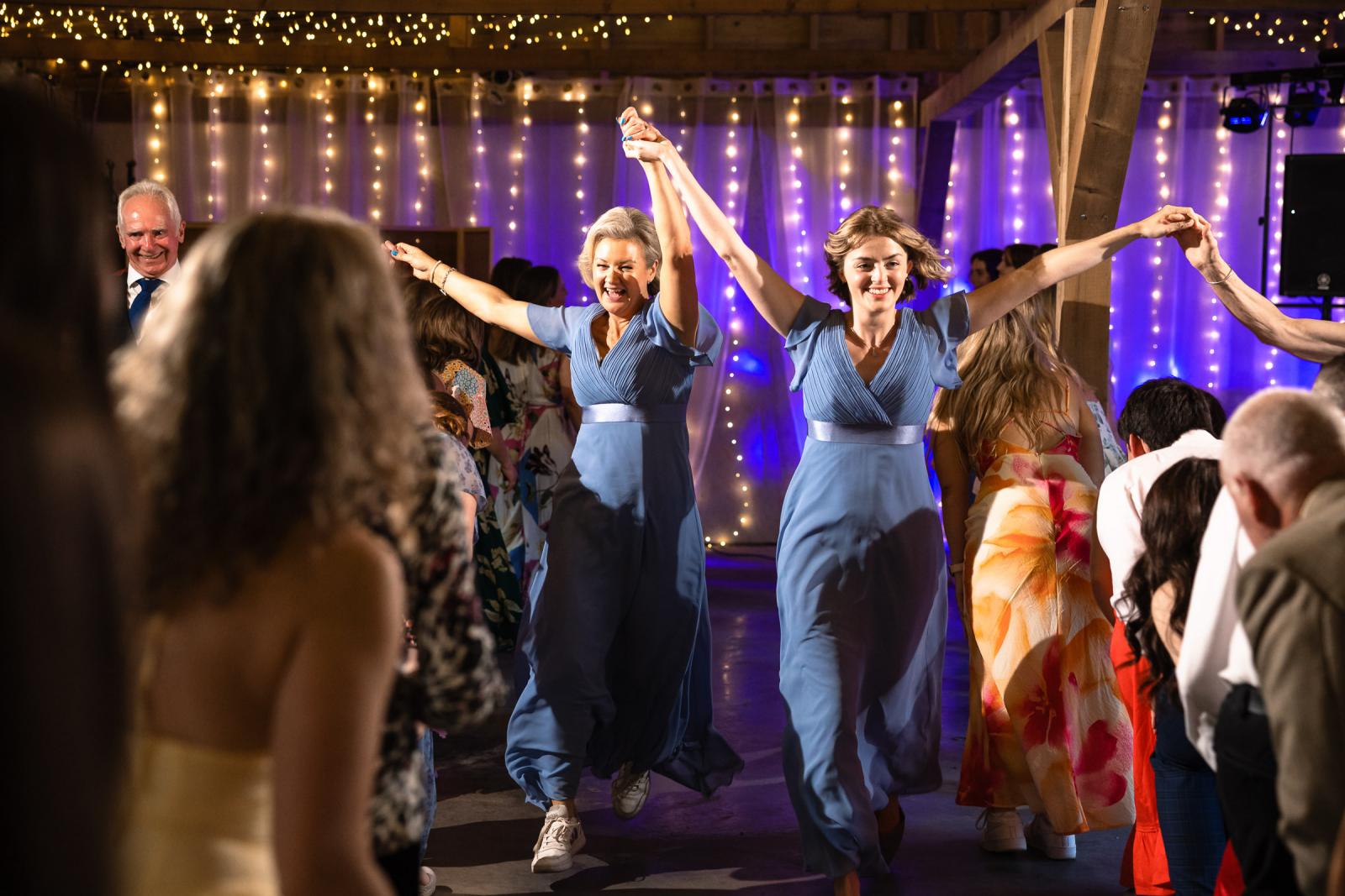 Bridesmaids in matching blue gowns joyfully raise their hands and dance during a traditional Scottish Ceilidh inside the illuminated reception barn.