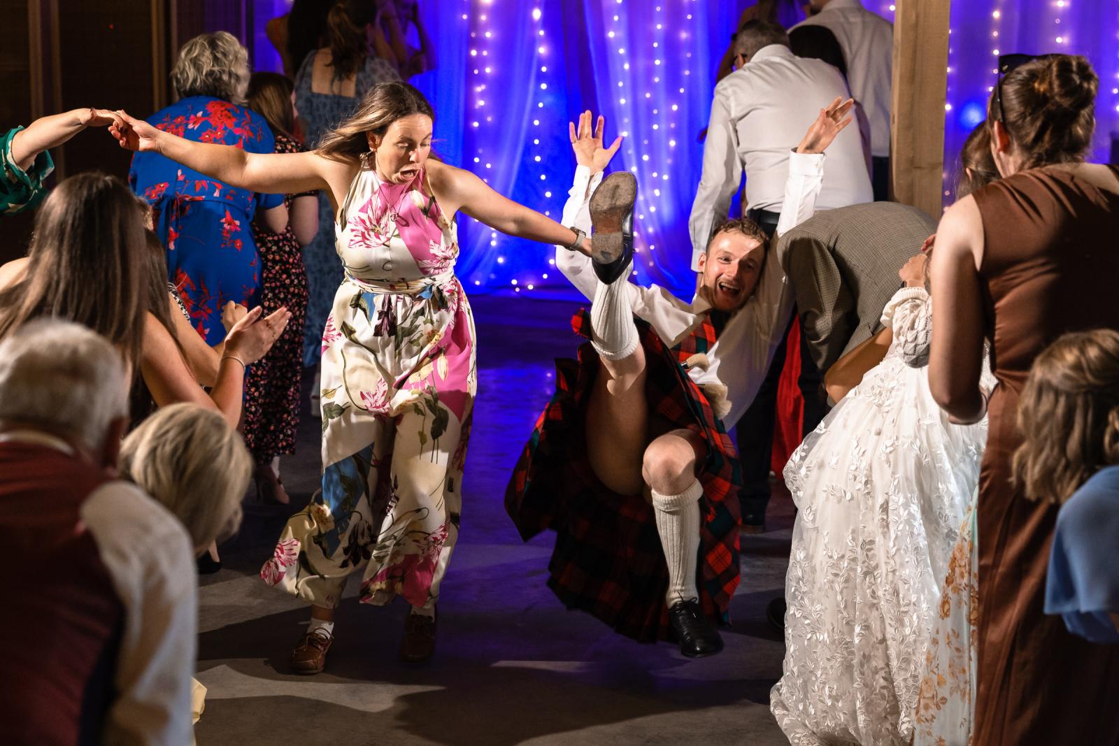 A hilarious, candid action shot of a wedding guest in a kilt falling backward onto the dance floor with his leg in the air during an energetic Ceilidh dance.