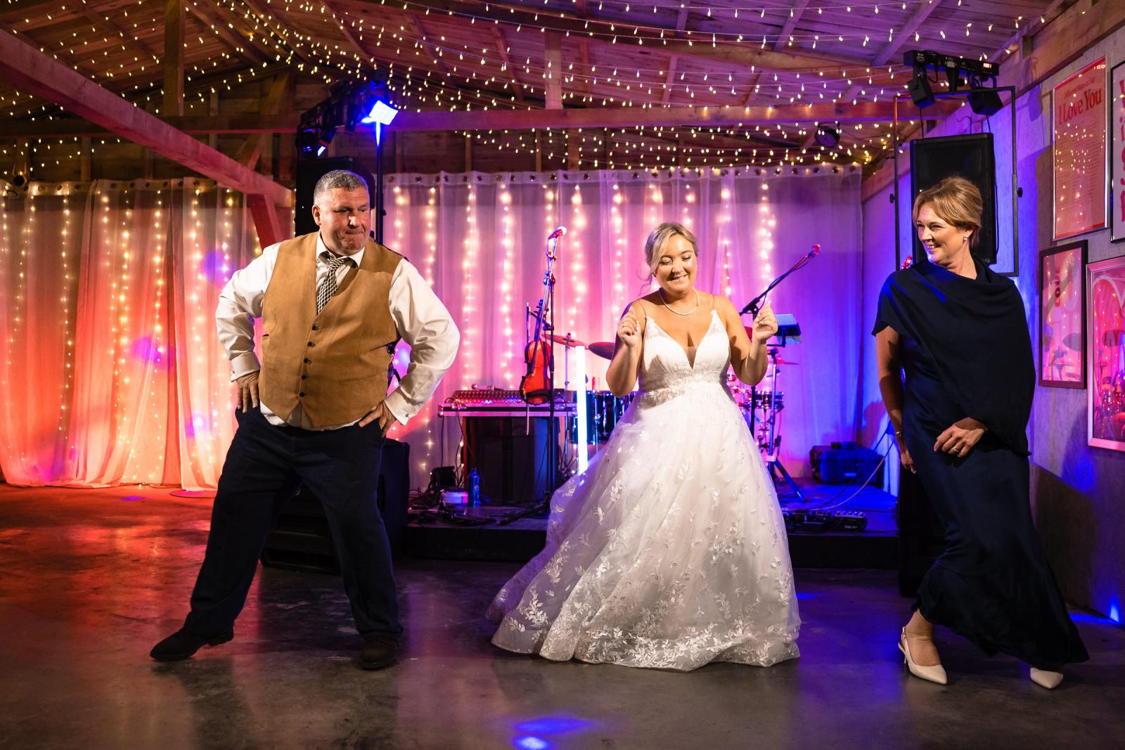 Bride Emma happily dances on the floor alongside two wedding guests, set against a backdrop of warm fairy lights and a stage setup.