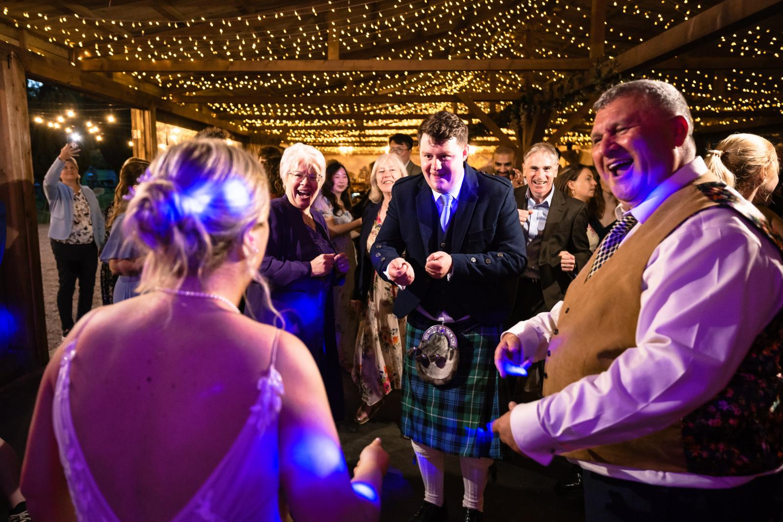 A lively, close-up shot of groom Hamish dancing in his traditional kilt alongside smiling wedding guests under a canopy of twinkling lights.