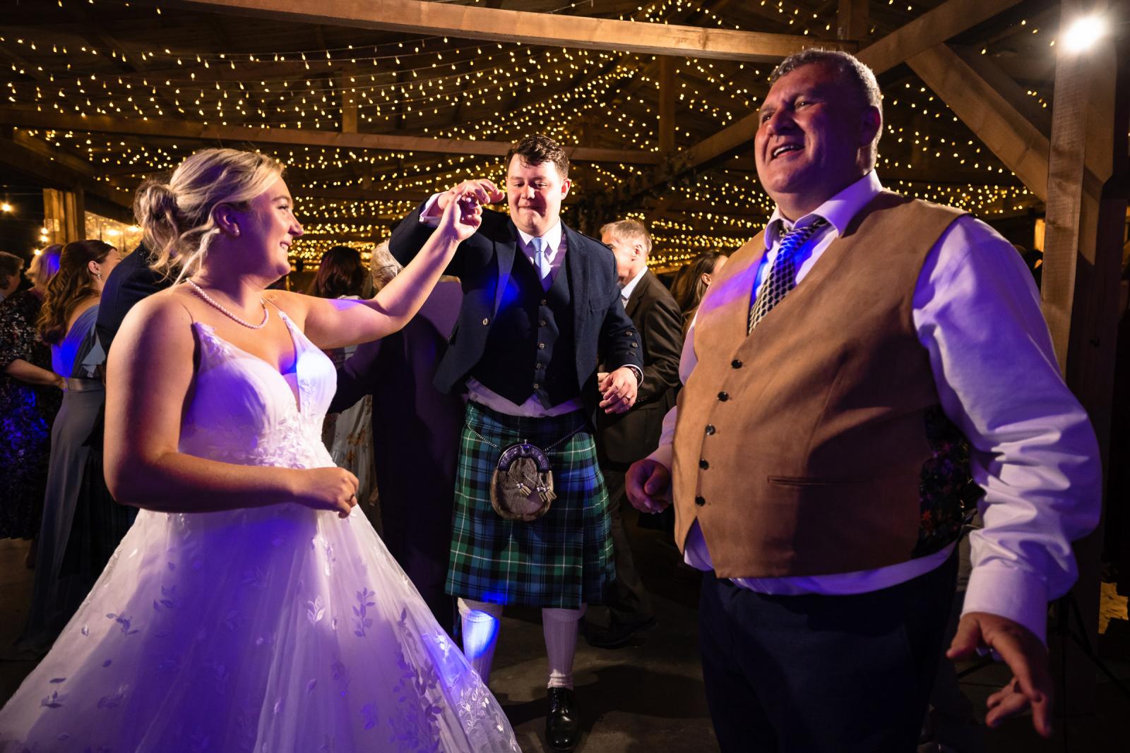 Bride Emma and groom Hamish smile at each other while holding hands and dancing on a crowded dance floor next to an enthusiastic guest.