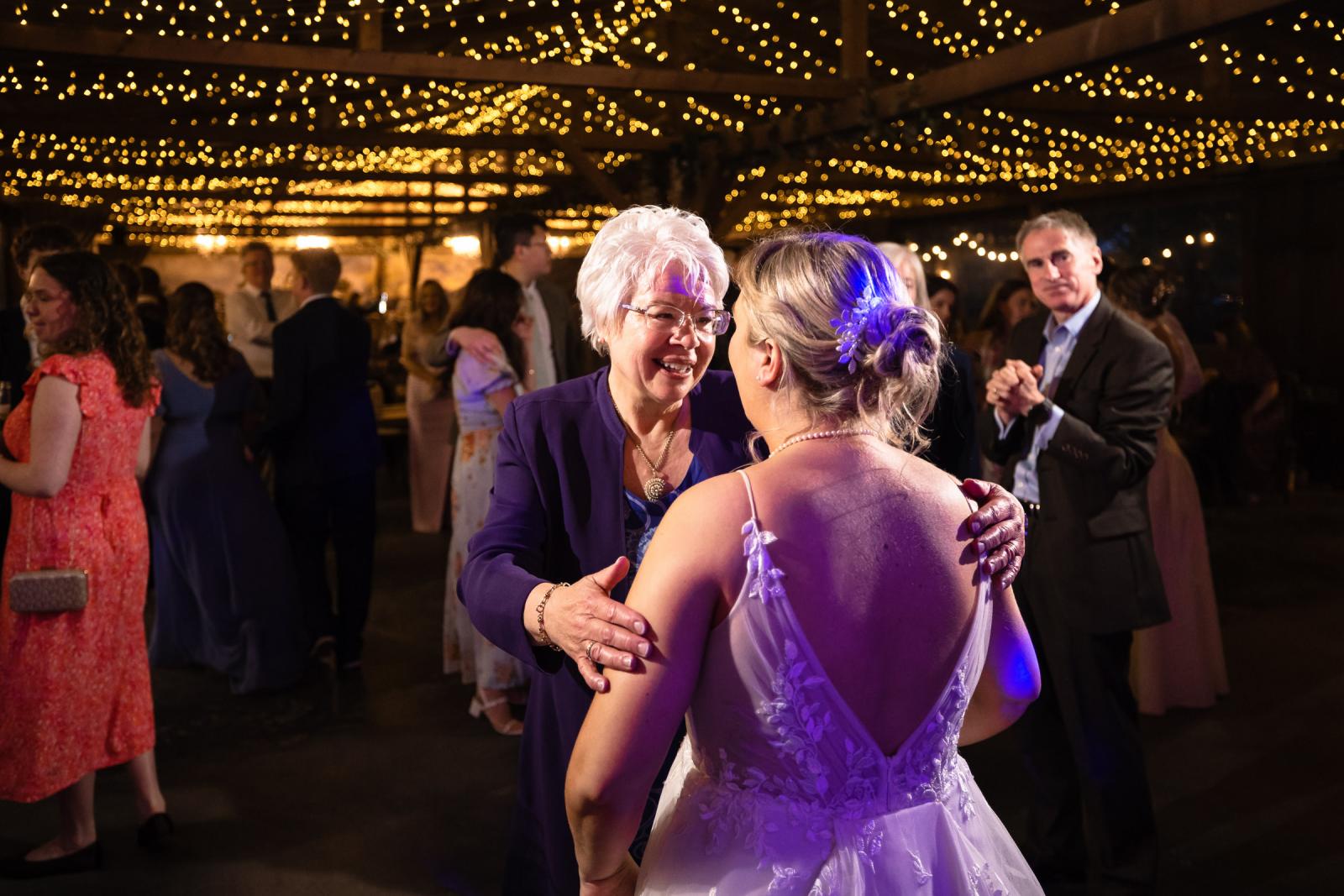 Mother of the groom in a purple jacket warmly embraces bride Emma on the lit dance floor as others look on.