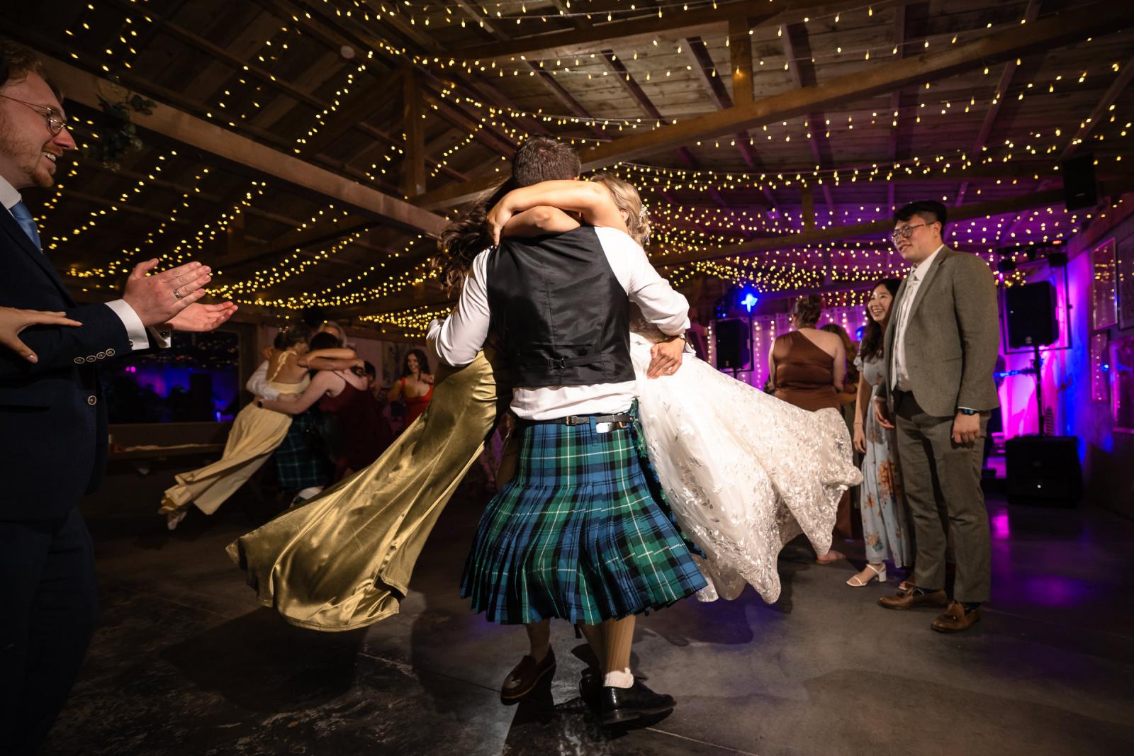 Groom Hamish in his Scottish kilt lifts a wedding guest into the air on the lively dance floor, surrounded by clapping and cheering onlookers.