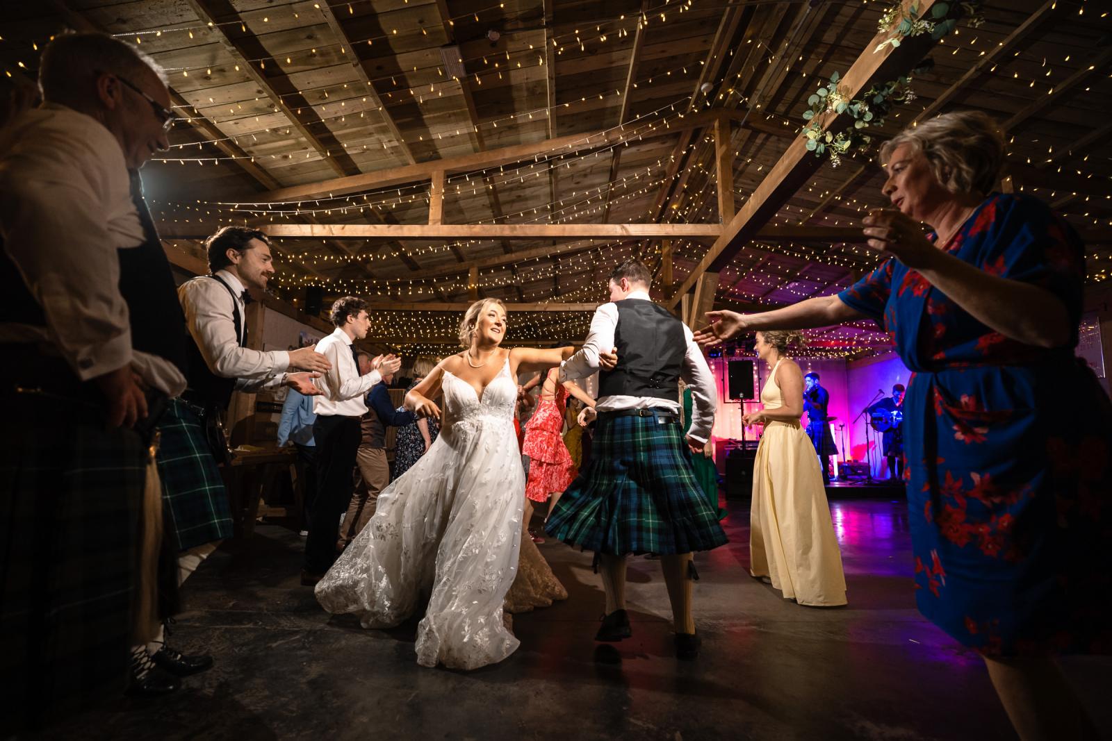 Bride Emma and groom Hamish dance energetically hand-in-hand in the center of a Ceilidh dance circle inside the warmly lit rustic barn.