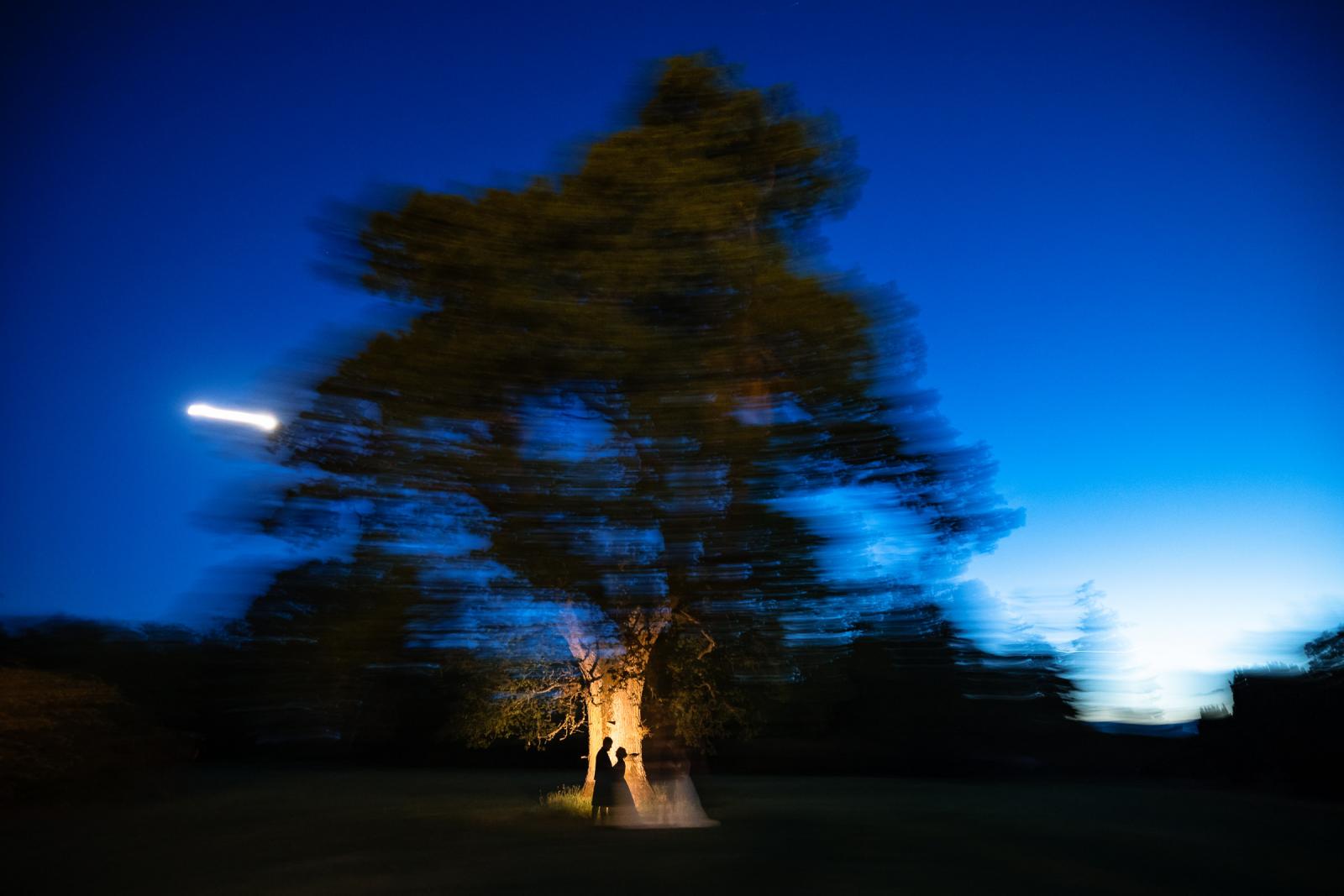 A creative, long-exposure nighttime photograph capturing the silhouette of a couple embracing under a beautifully illuminated tree against a deep blue sky.