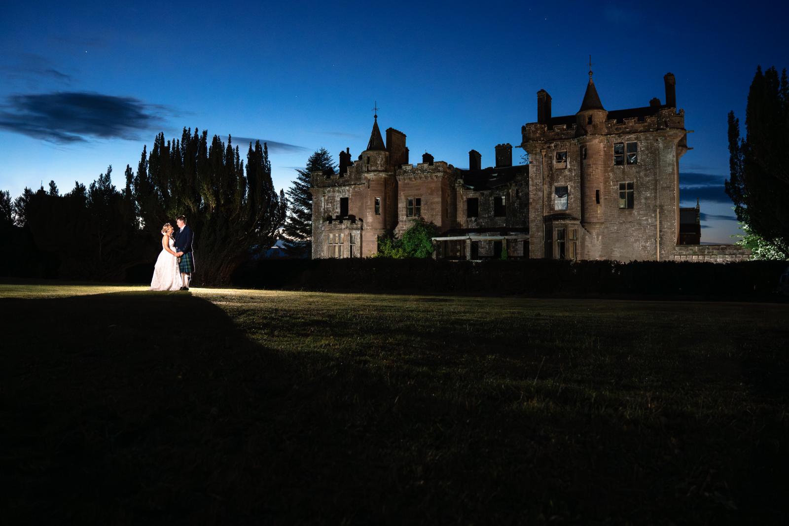 A striking night portrait of Emma and Hamish standing hand-in-hand on the dark lawn of Culdees Castle Estate, with the historic stone castle lit up behind them.