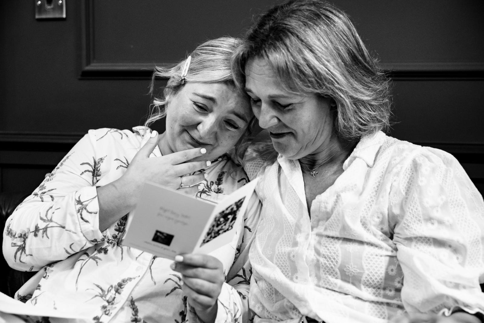 Black and white emotional wedding photograph of Emma and her mother crying tears of joy while reading a handwritten card together in a close embrace.