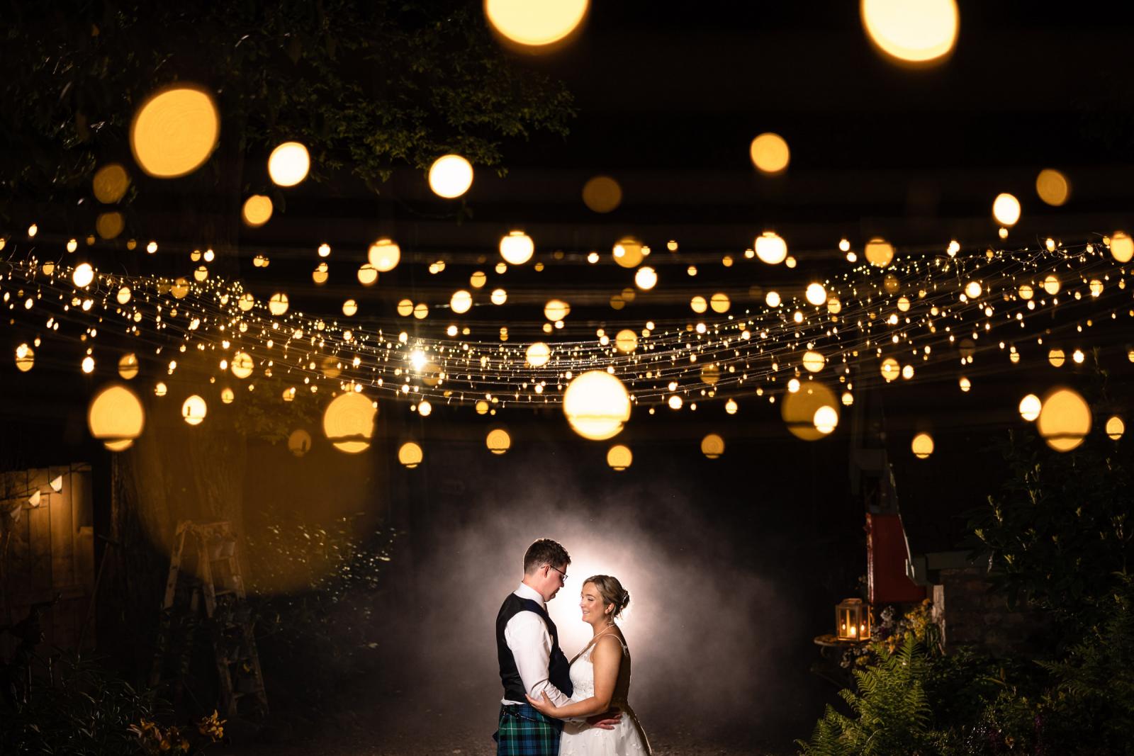 A romantic, atmospheric night portrait of Emma and Hamish holding each other close amidst a dramatic haze, beautifully framed by a canopy of warm, glowing fairy lights.