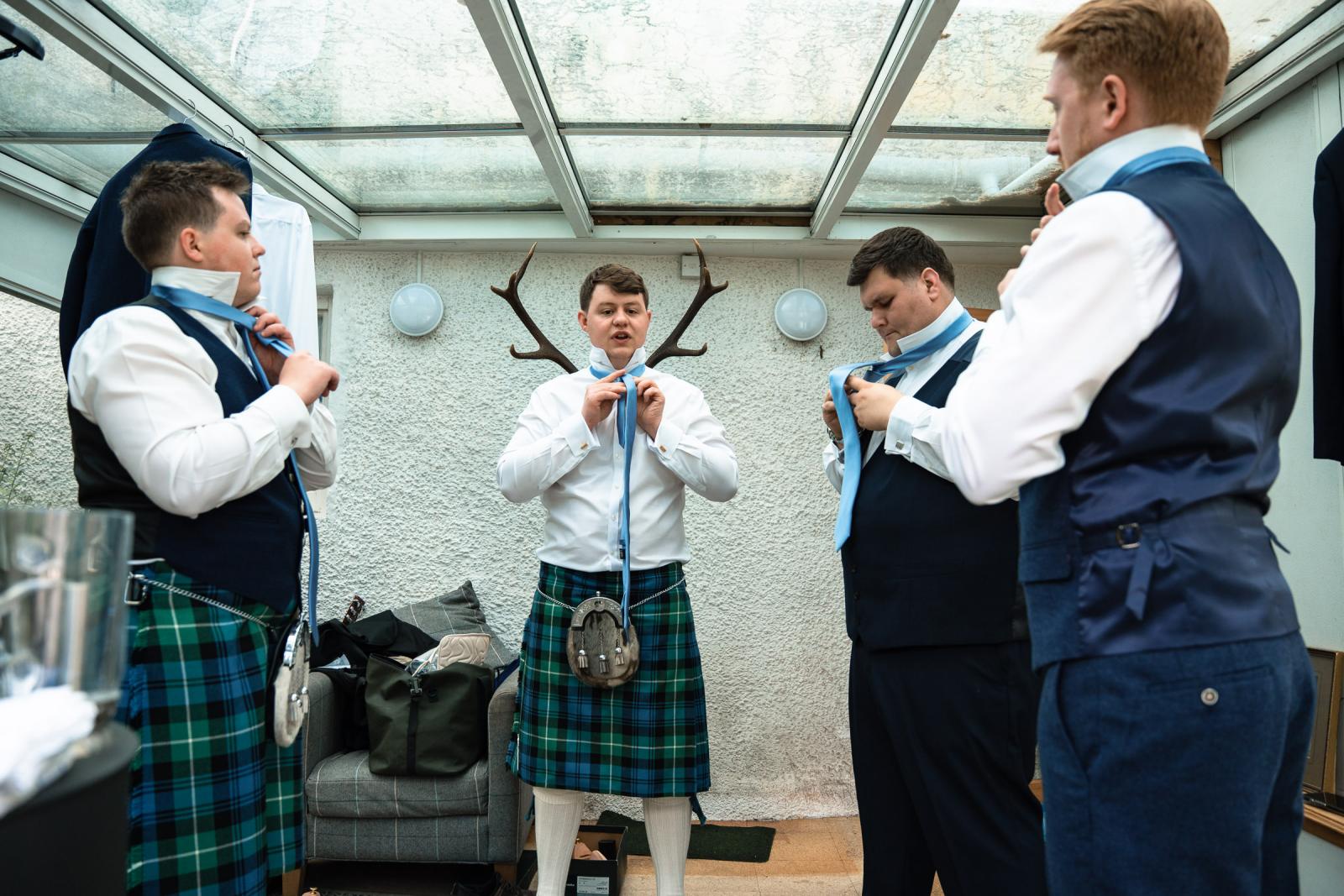 The groom Hamish and his three groomsmen standing in a row in a conservatory, helping each other tie their light blue silk ties with their kilts on.