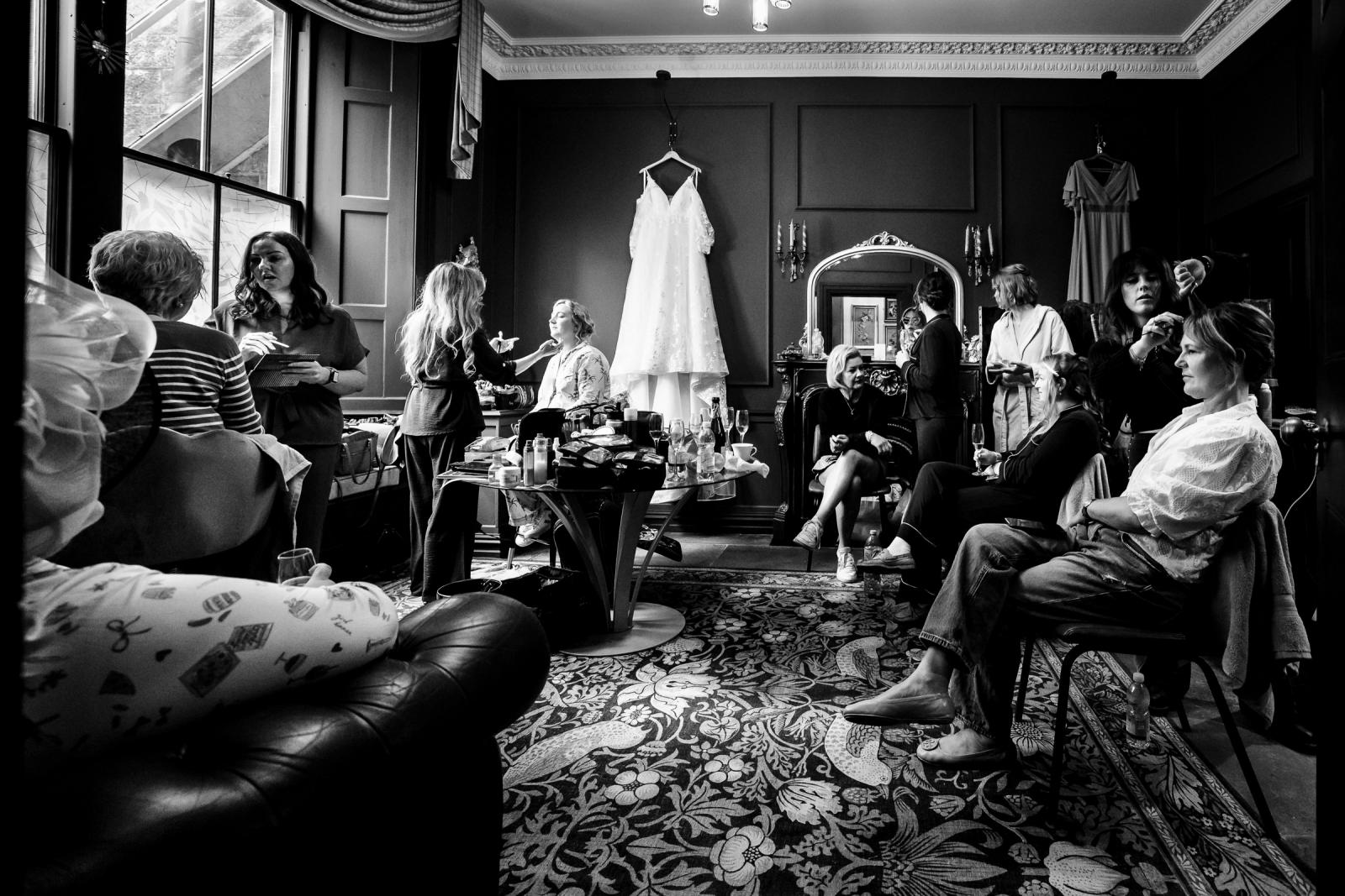 Wide black and white documentary photograph capturing the busy bridal preparation room at Culdees Castle with bridesmaids and family members getting hair and makeup done.