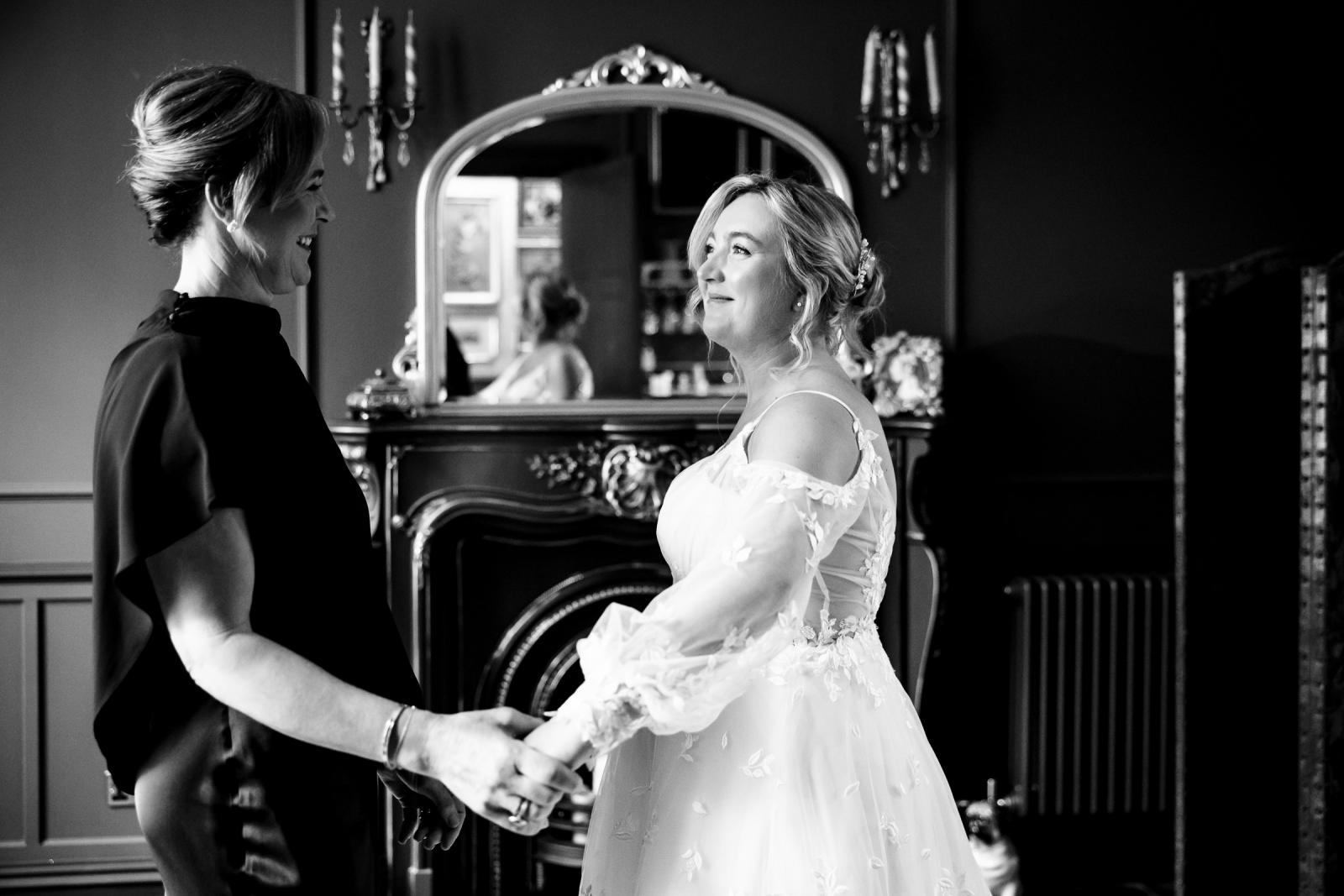 Emotional black and white portrait of bride Emma in her wedding gown holding hands and sharing a tender look with her mother in front of a castle fireplace.