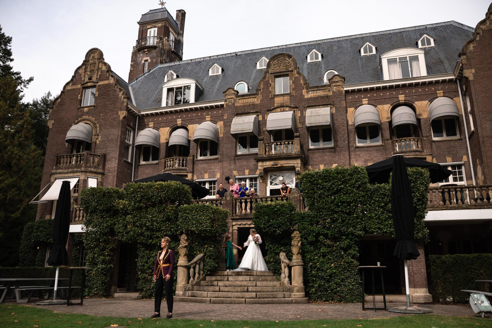 De first look: twee bruiden zien elkaar voor het eerst bij Kasteel De Hooge Vuursche terwijl familie toekijkt vanaf het bordes.
