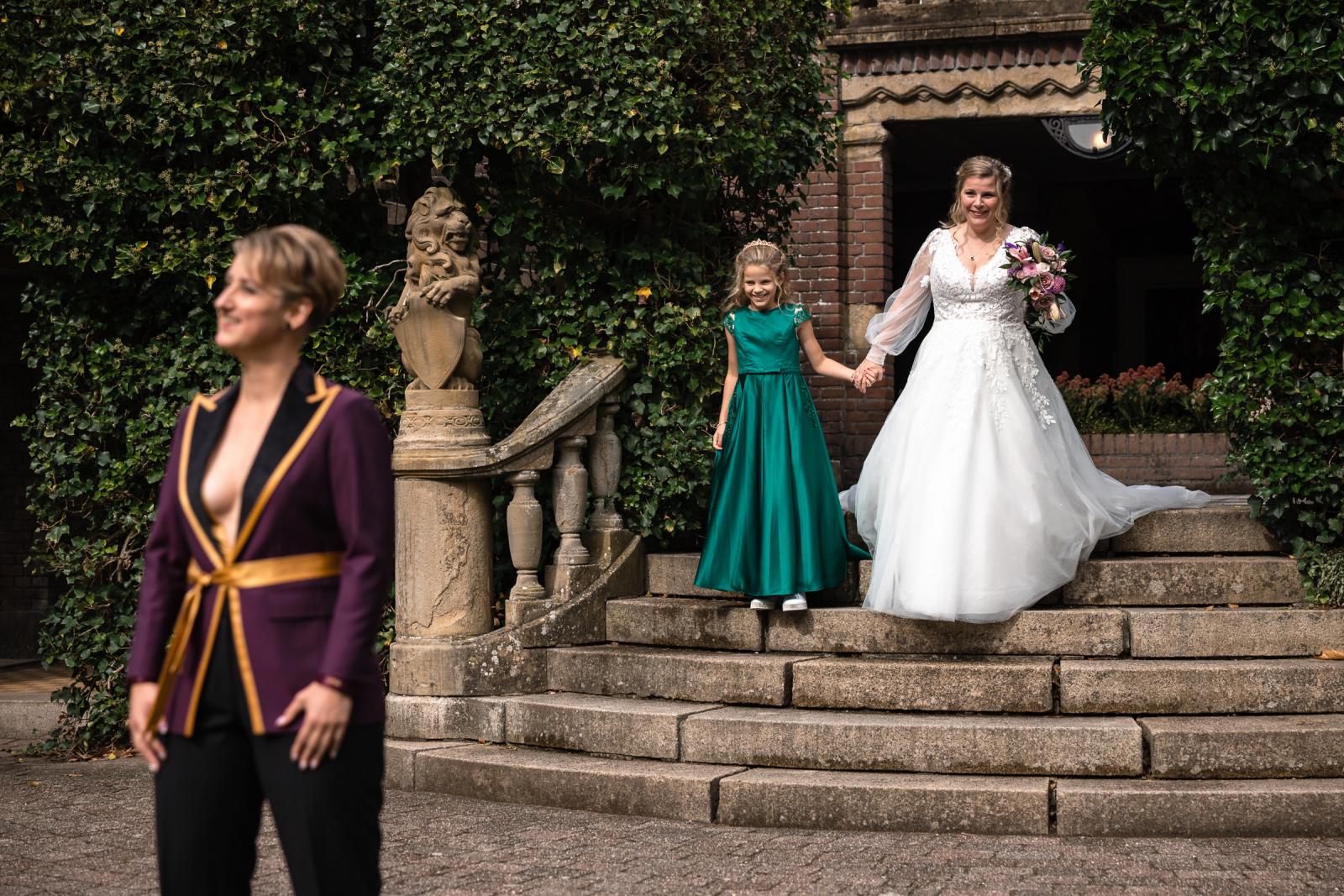 De bruid in witte jurk loopt hand in hand met haar dochter de trap af voor de first look bij Kasteel De Hooge Vuursche.