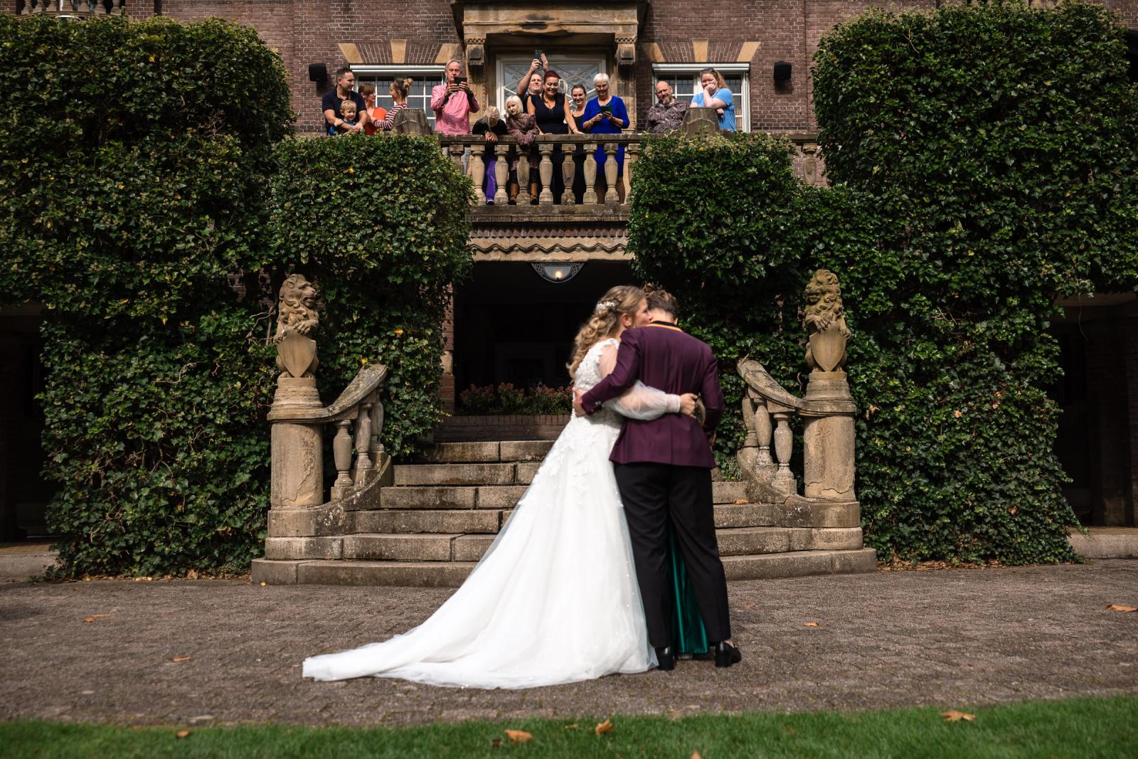 Twee bruiden omhelzen elkaar tijdens de first look terwijl familie applaudisseert vanaf het balkon van Kasteel De Hooge Vuursche.