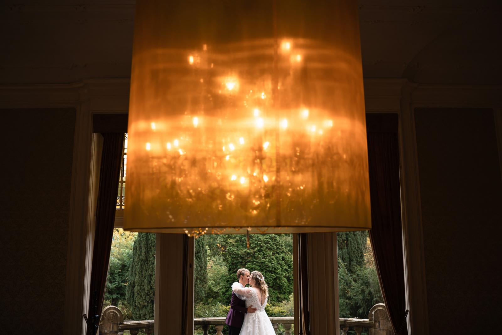 Intiem moment van de twee bruiden die elkaar omhelzen onder een grote kroonluchter in het kasteel.