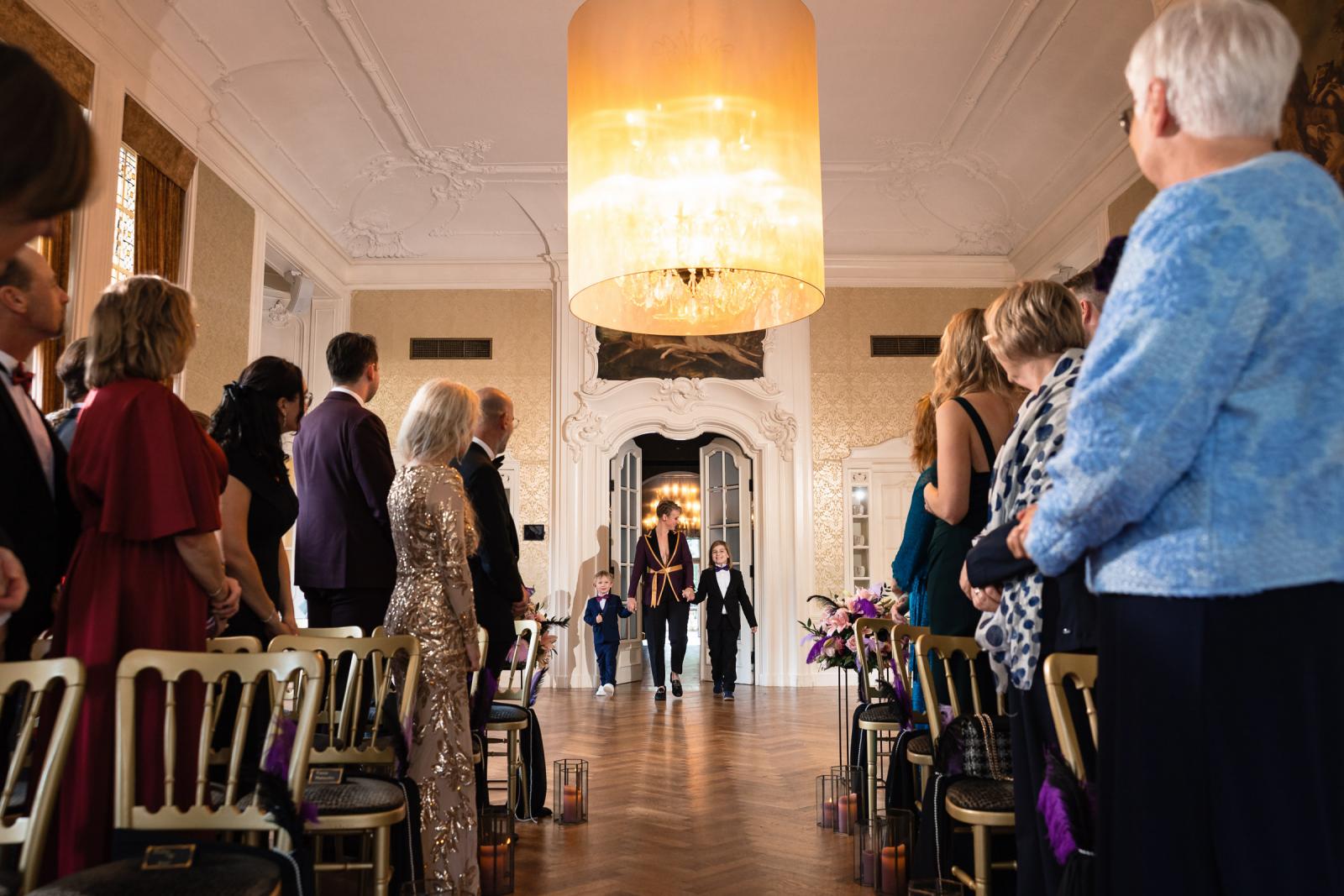 De ceremonie begint: n van de bruiden loopt met twee kinderen de trouwzaal binnen terwijl gasten opstaan.