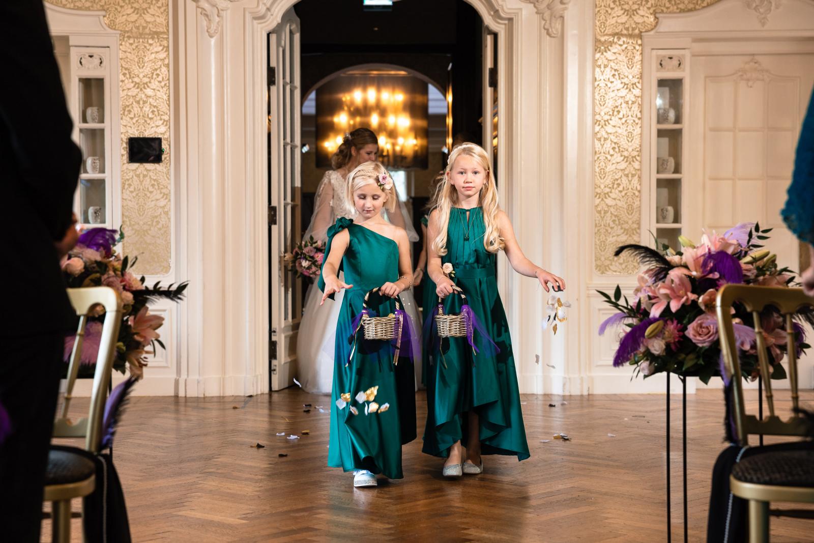 Twee bloemenmeisjes in groene jurken strooien bloemblaadjes in het gangpad terwijl de bruid op de achtergrond verschijnt.