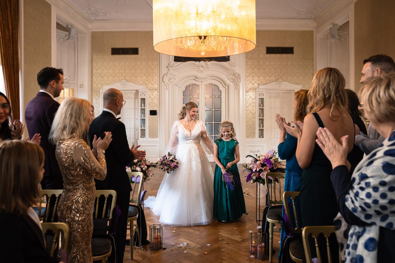 De bruid loopt hand in hand met haar dochter het gangpad in terwijl gasten applaudisseren in de trouwzaal van Kasteel De Hooge Vuursche.
