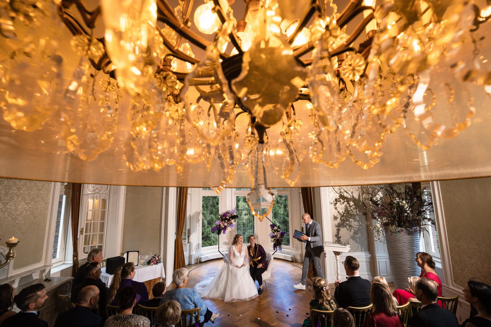Overzichtsfoto van de ceremonie met de twee bruiden onder een grote kroonluchter in de stijlvolle trouwzaal van het kasteel.