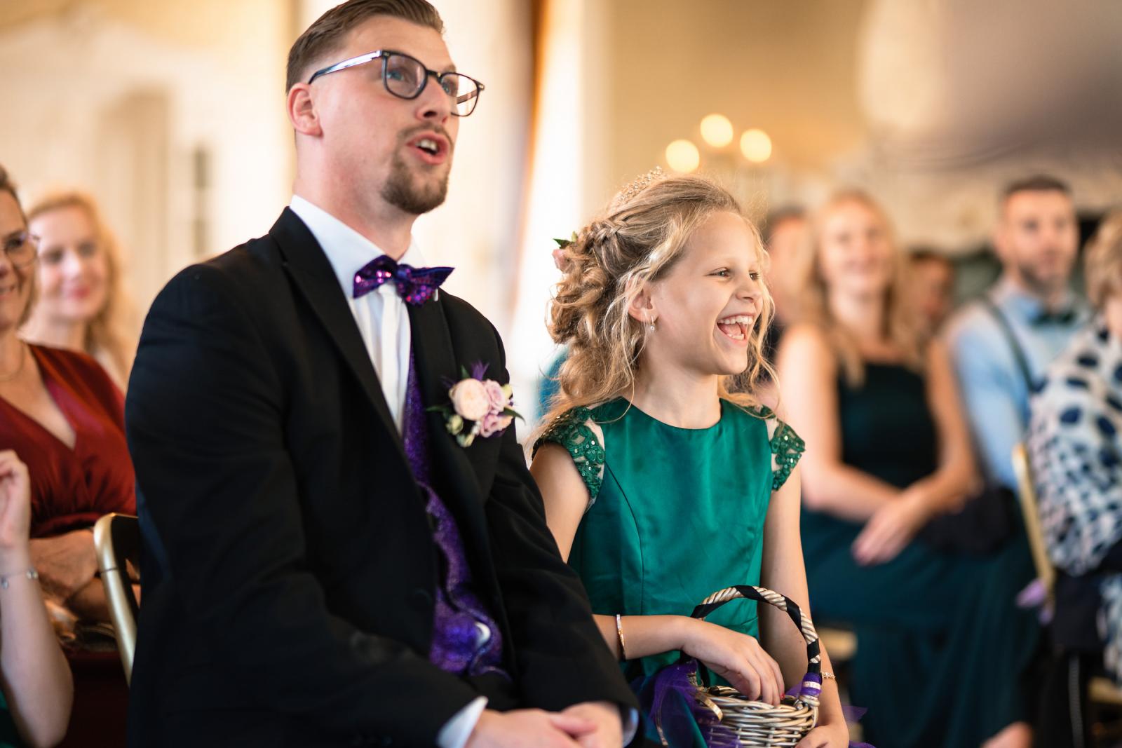De dochter van de bruiden lacht uitbundig tijdens de ceremonie, zittend naast een gast in pak met paarse details.