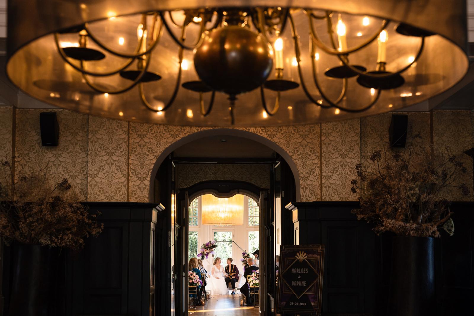 Blik door de deuropening richting de ceremonie, waar de twee bruiden zitten onder een bloemrijke boog in de trouwzaal van het kasteel.