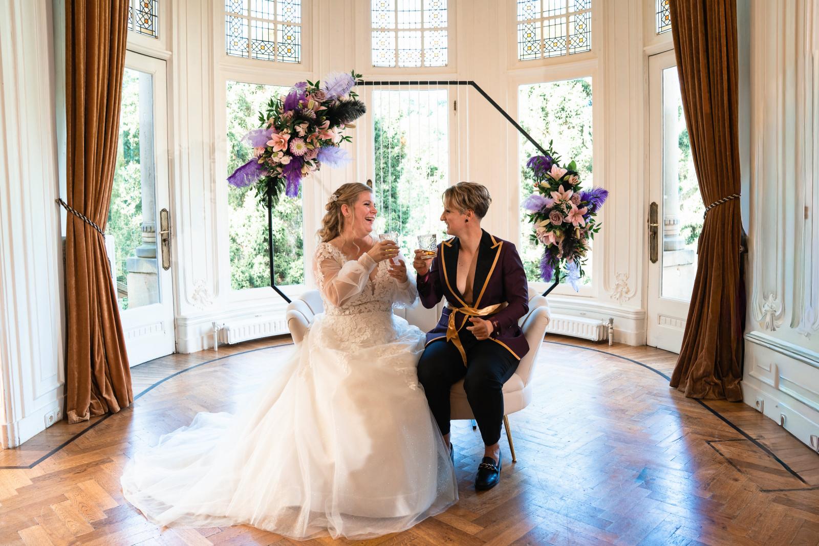 De twee bruiden proosten op hun ceremonie, zittend voor de bloemstukken in zachte pastelkleuren.