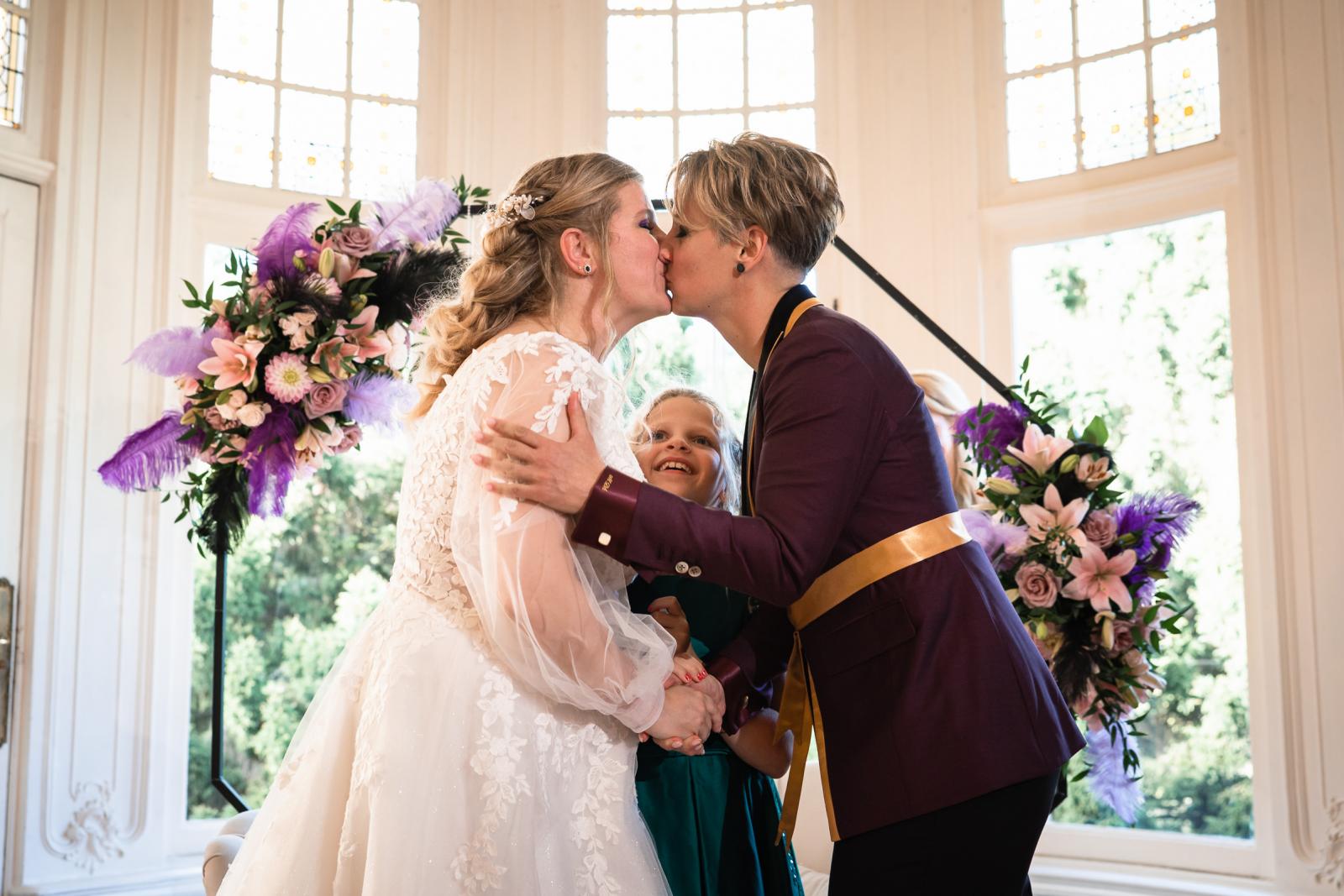 De twee bruiden delen hun eerste kus na het jawoord terwijl hun dochter blij tussen hen in staat.
