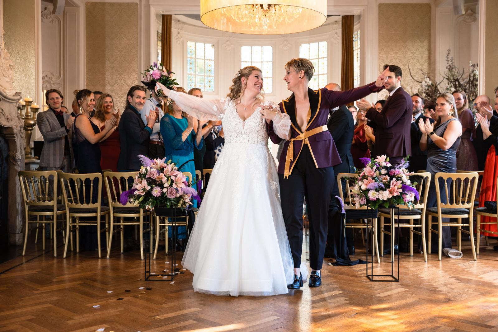 De bruiden lopen hand in hand de trouwzaal uit terwijl gasten klappen en bloemen strooien.