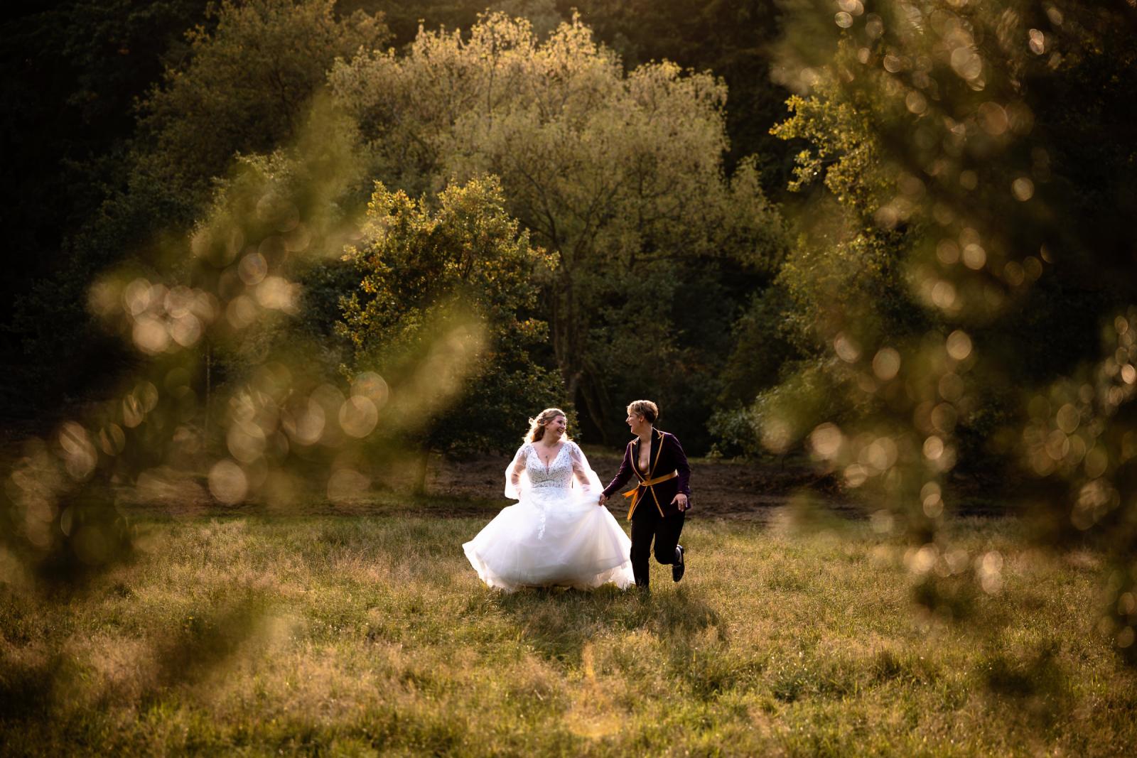 De twee bruiden rennen lachend hand in hand door een veld bij zonsondergang.