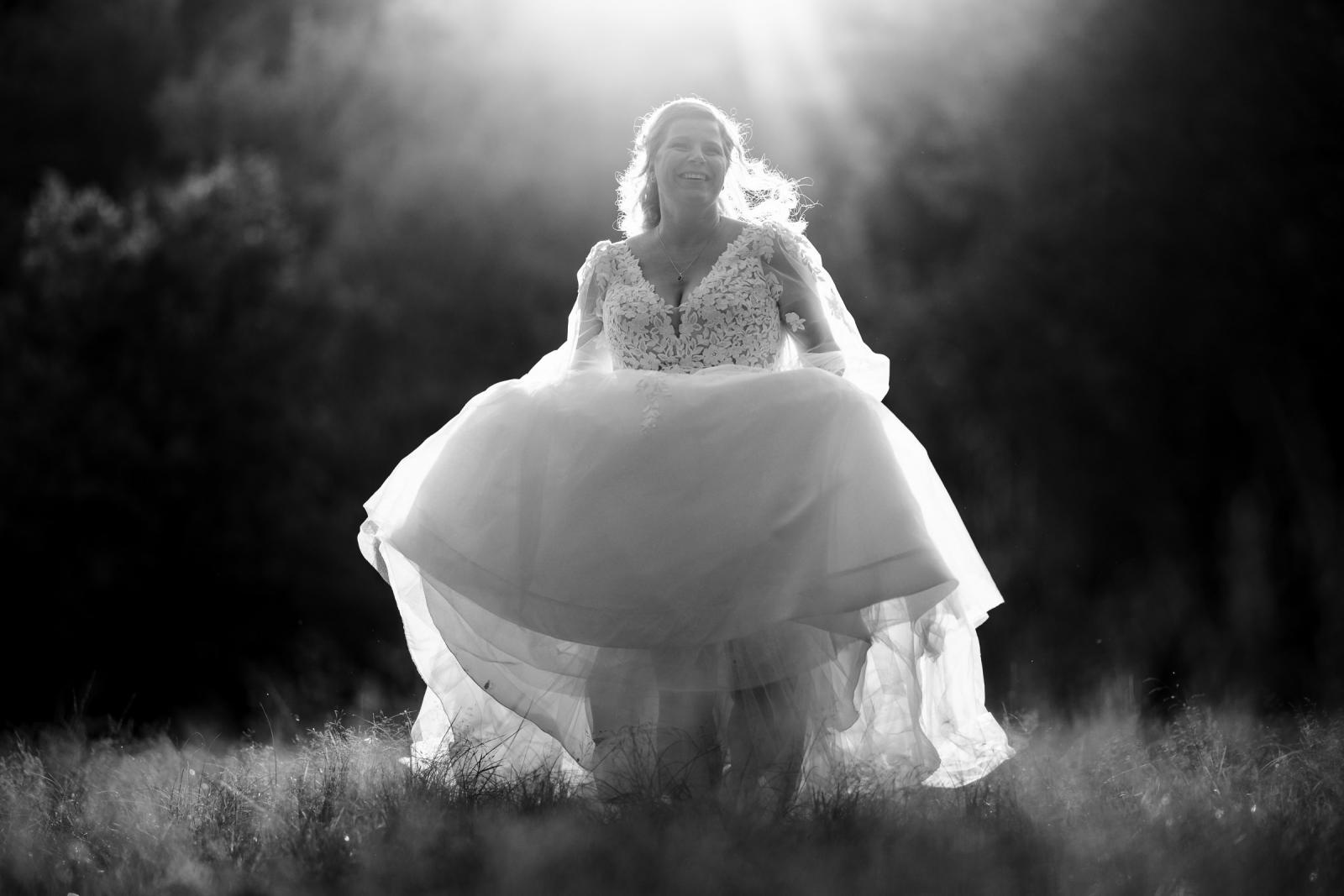 Zwart-wit foto van de bruid die haar jurk optilt terwijl ze door het gras loopt in het tegenlicht.