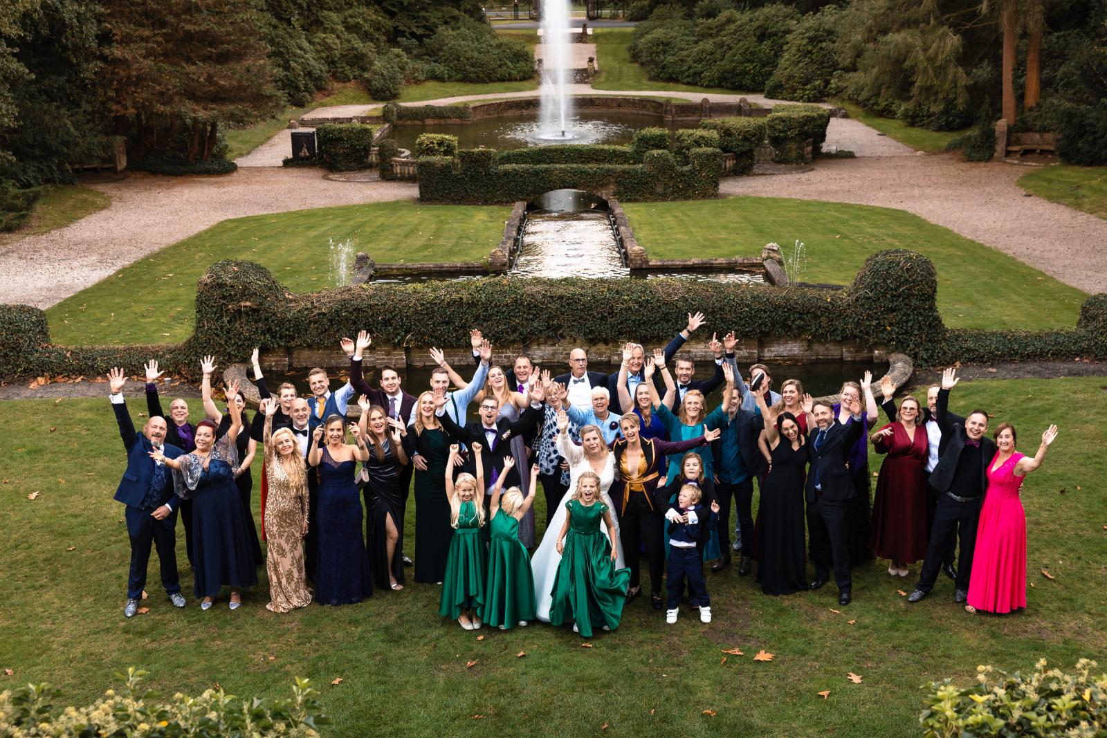 Groepsfoto van alle bruiloftsgasten in de tuin van Kasteel De Hooge Vuursche met de fontein op de achtergrond.