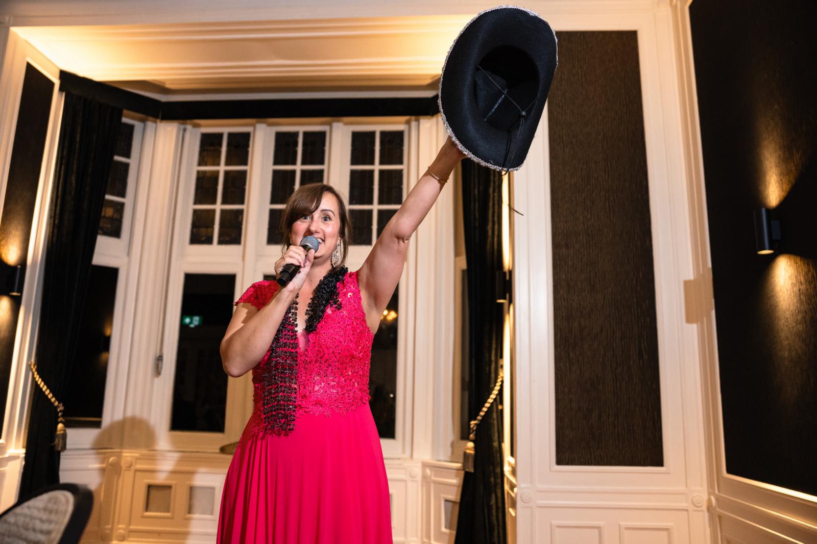 Gaste in roze jurk kondigt een verrassingsoptreden aan met een zwarte hoed in haar hand.