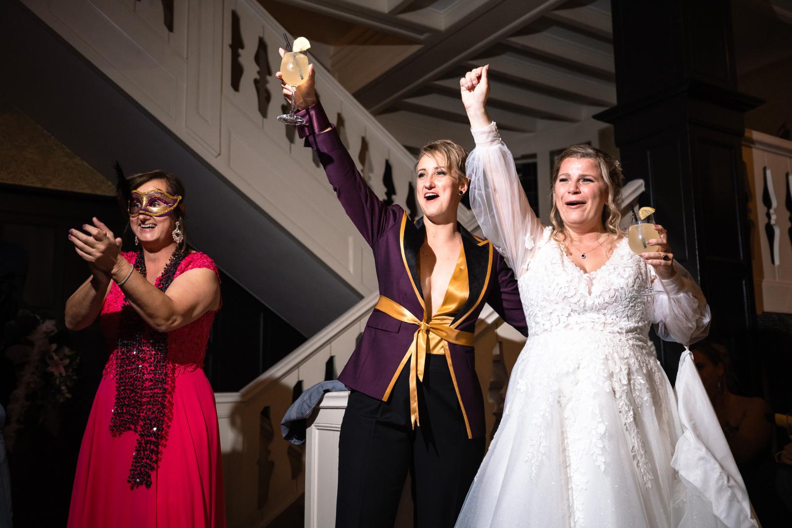 De bruiden proosten en juichen samen tijdens het avondfeest, omringd door gasten in feestmaskers.