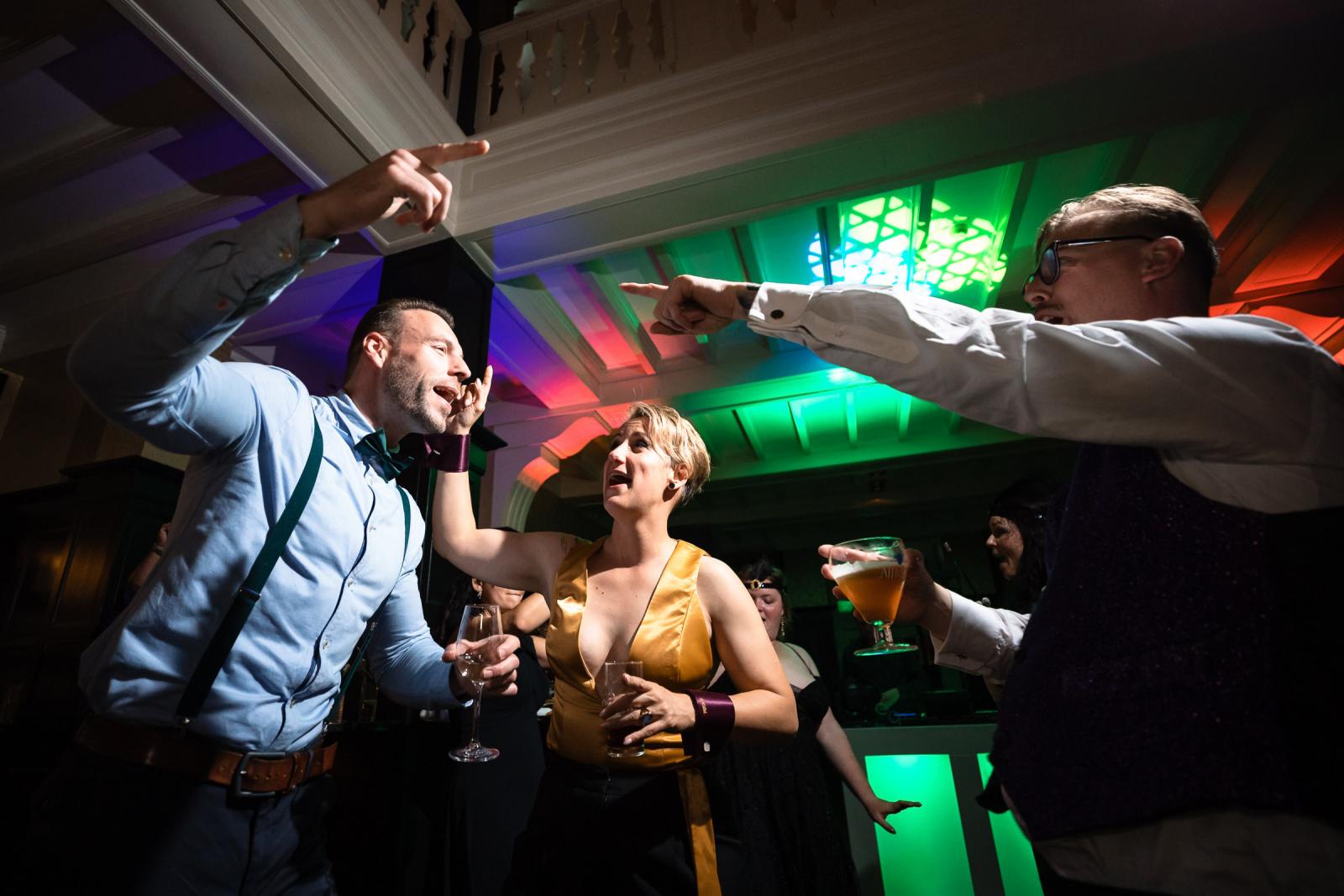 De bruid in goud en een gast zingen enthousiast mee op de dansvloer onder kleurrijke feestverlichting.