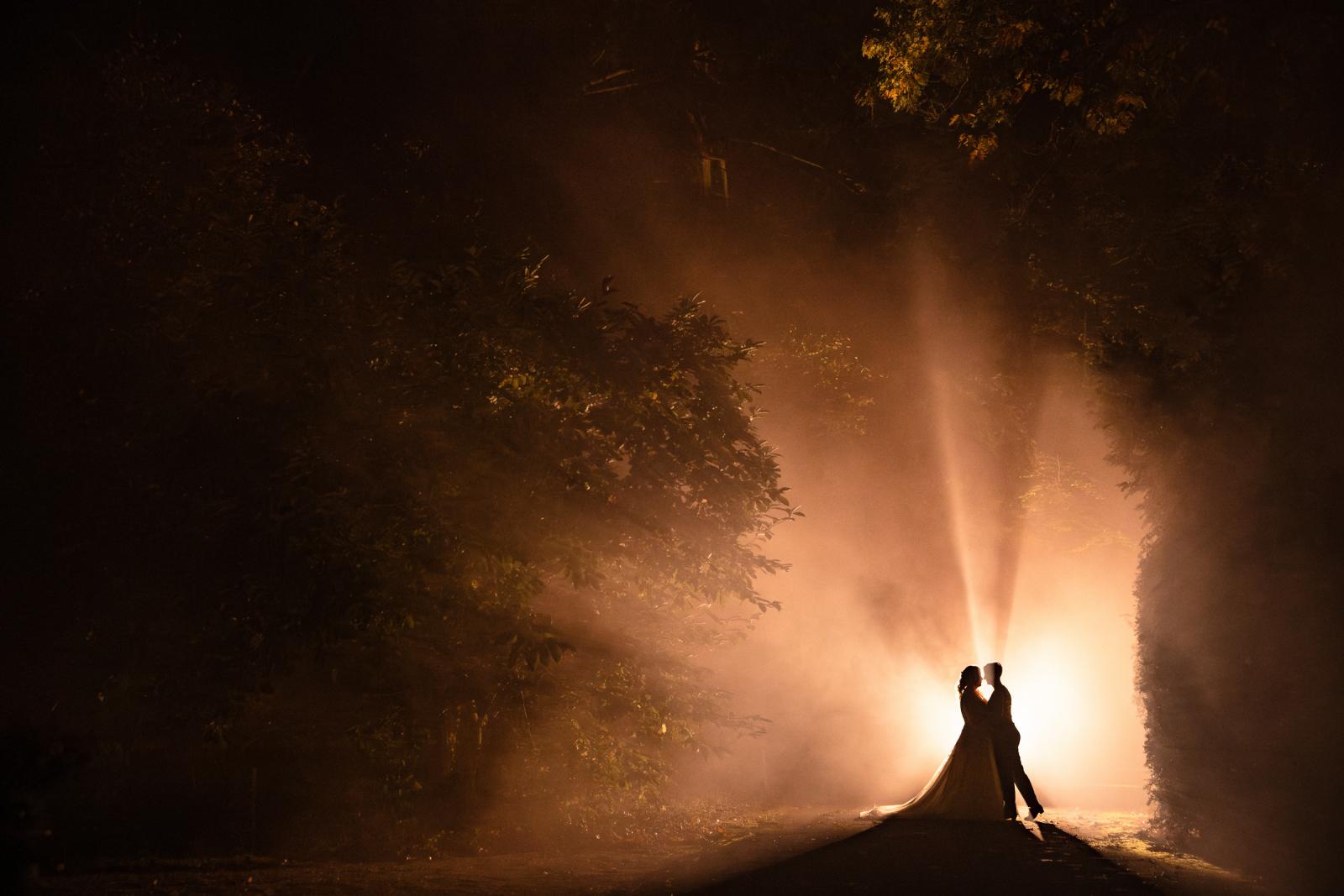 Silhouet van twee bruiden in een intieme omhelzing bij avondlicht, omgeven door mist en bomen.