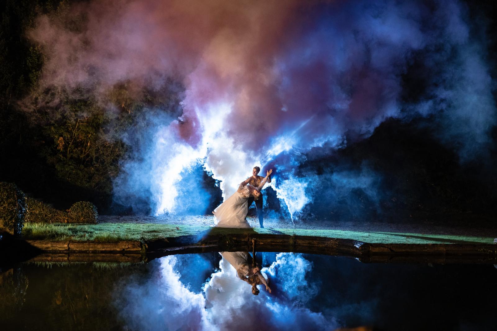 De bruiden dansen bij het water terwijl gekleurde rook zich weerspiegelt in de vijver, een magisch moment tijdens de avondshoot.