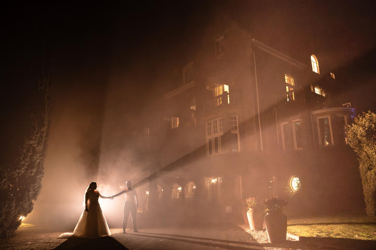 Romantische avondfoto van de bruiden hand in hand voor het verlichte kasteel, badend in warme nevel en lichtstralen.