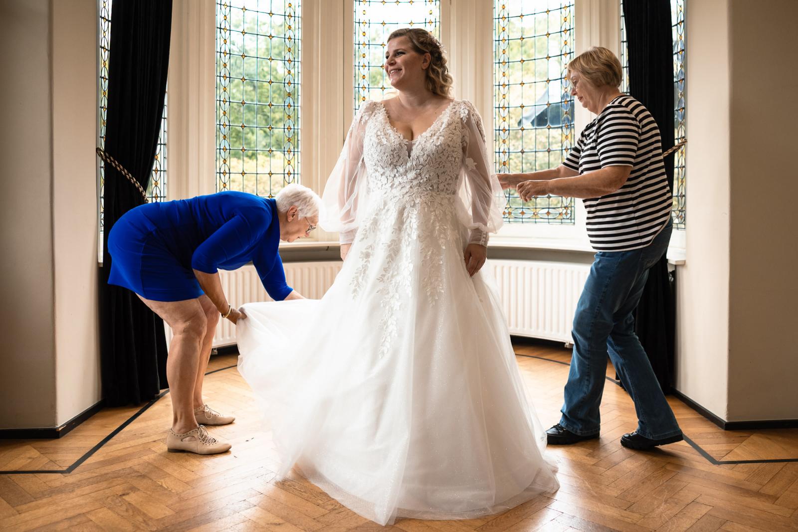 De bruid lacht terwijl haar jurk wordt rechtgetrokken door haar moeder in een lichte kamer met glas-in-loodramen.
