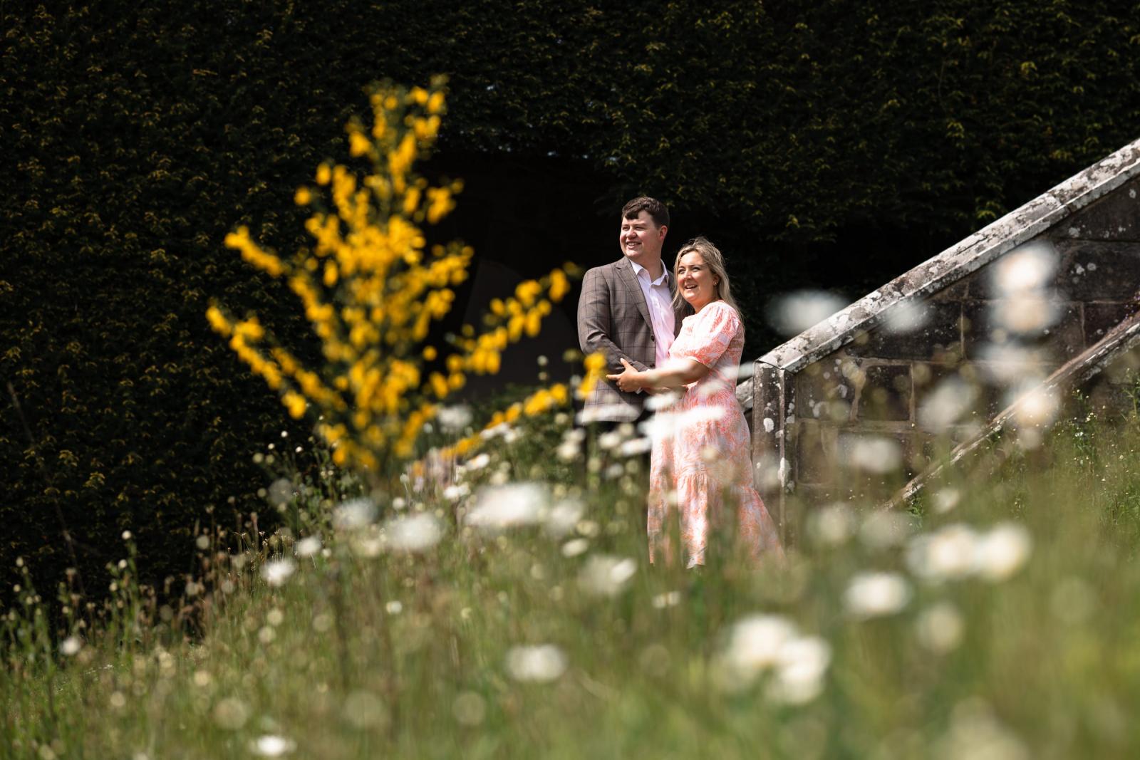 Emma and Hamish smiling together behind a field of white and yellow wildflowers in the castle garden