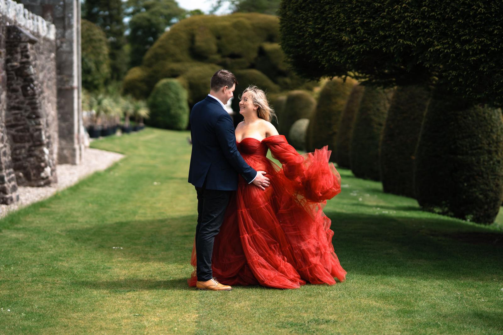 Editorial portrait of Emma in a flowing red dress and Hamish in a suit in the gardens of Drummond Castle