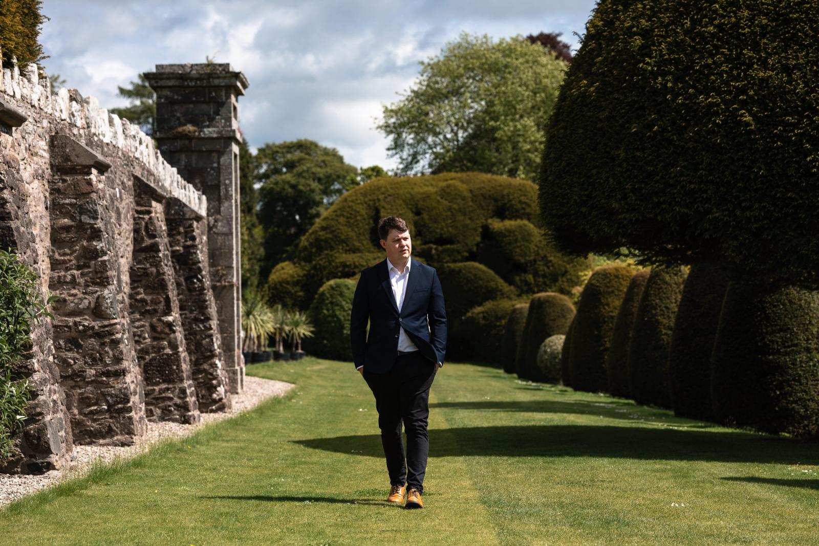 Hamish walking solo across a manicured lawn alongside a historic stone wall in Scotland