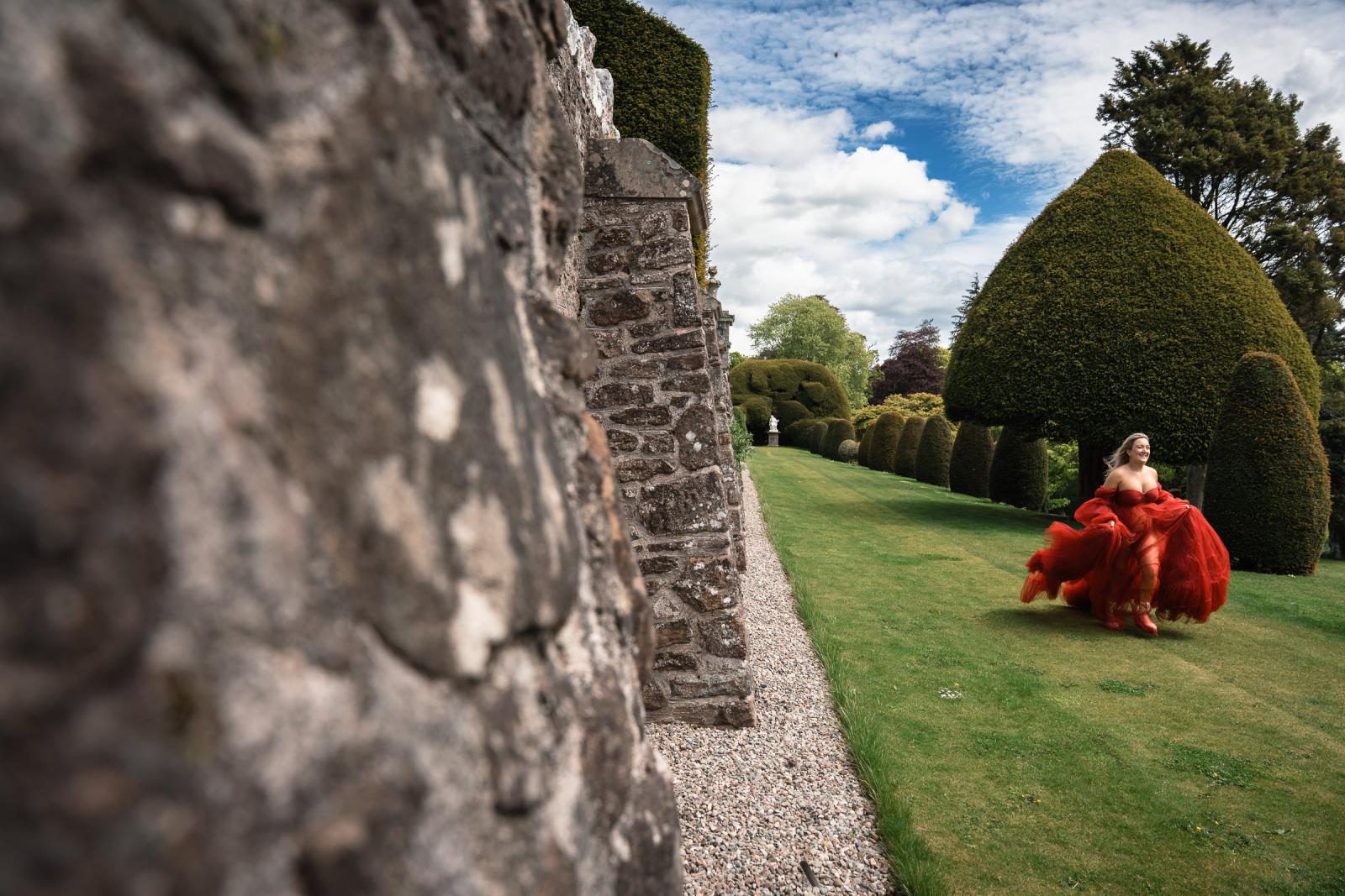 Emma running joyfully in her red dress across the lawn next to monumental hedges and stone walls